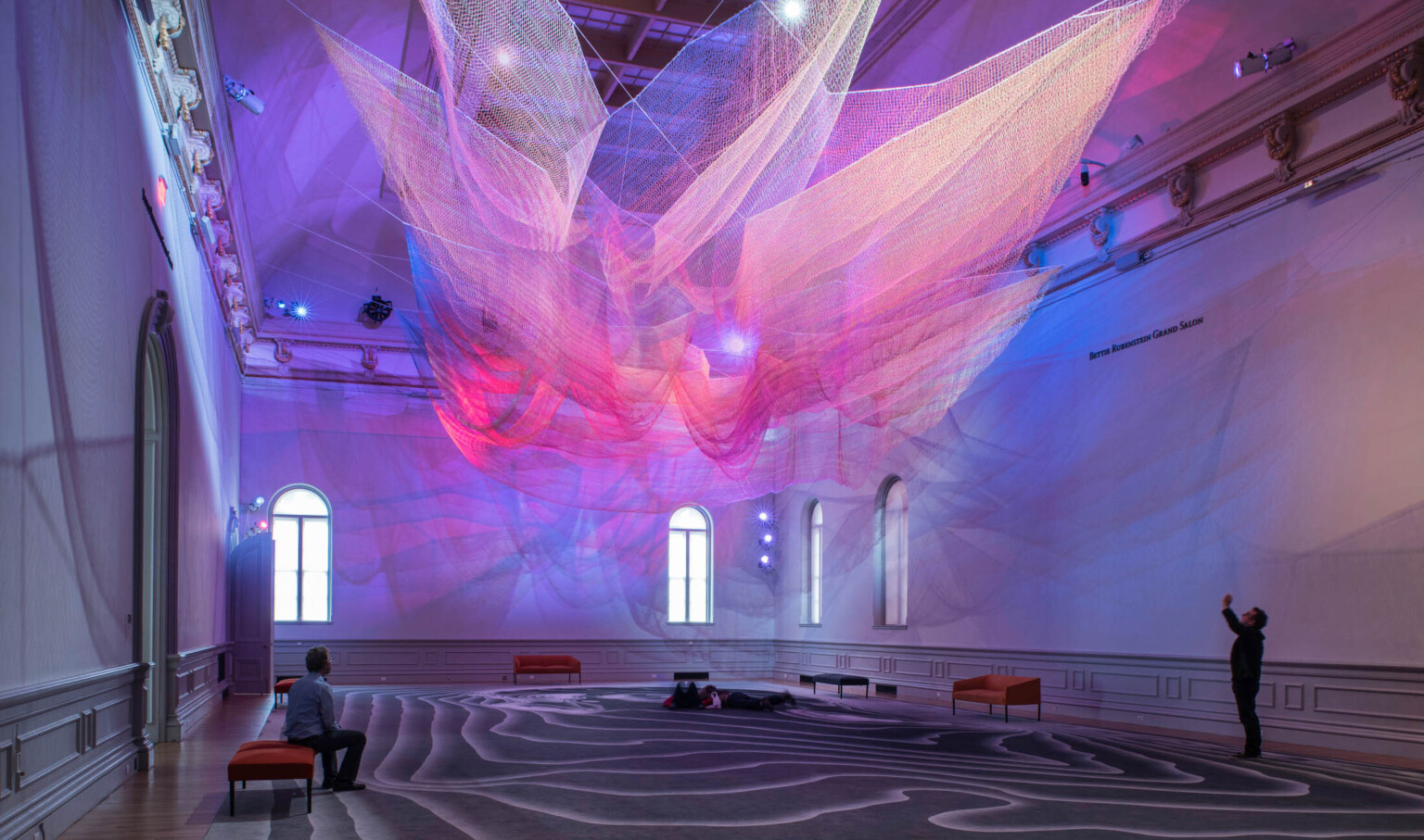 Colorful abstract art installation with pink and purple light on layers of sheer netting with shadows and light on white wall