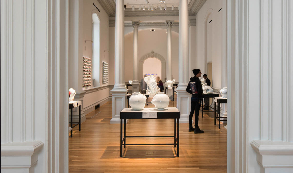 white art gallery with tall pillars displaying pottery on dark tables, framed by crown molding around doorway