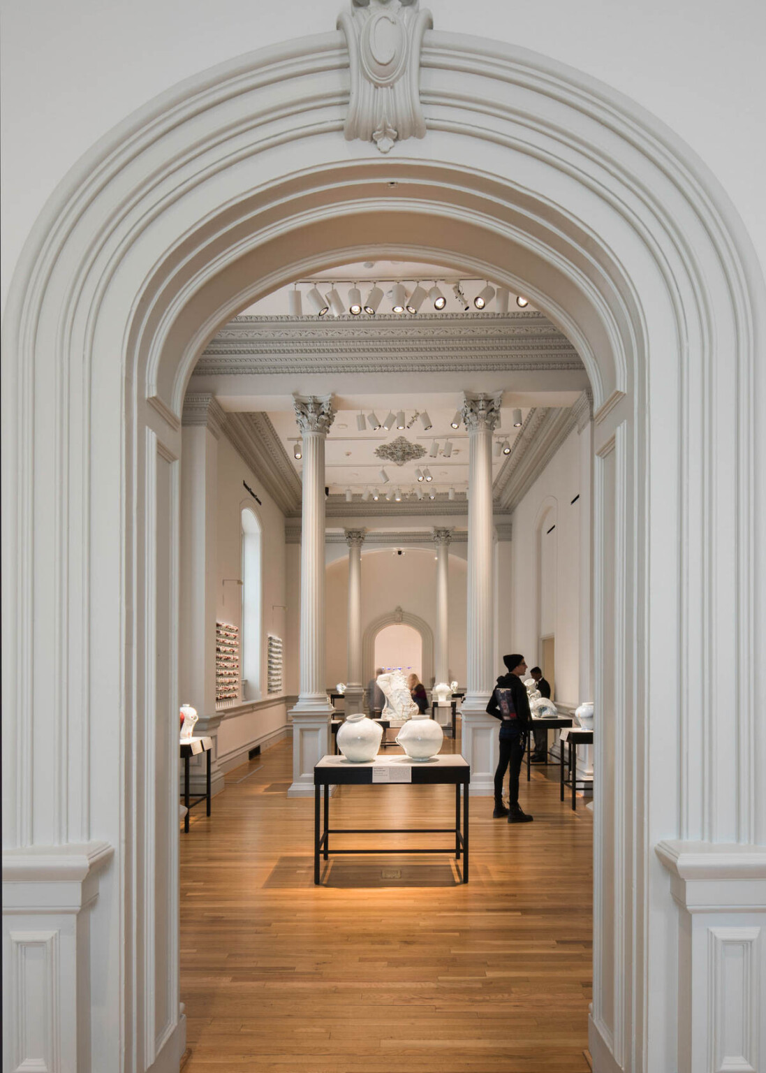 white art gallery with tall pillars displaying pottery on dark tables, framed by crown molding around doorway