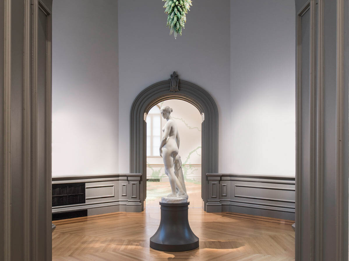 Marble sculpture of a woman on a black pedestal in the center of a small room, framed in front and behind by molded door frame