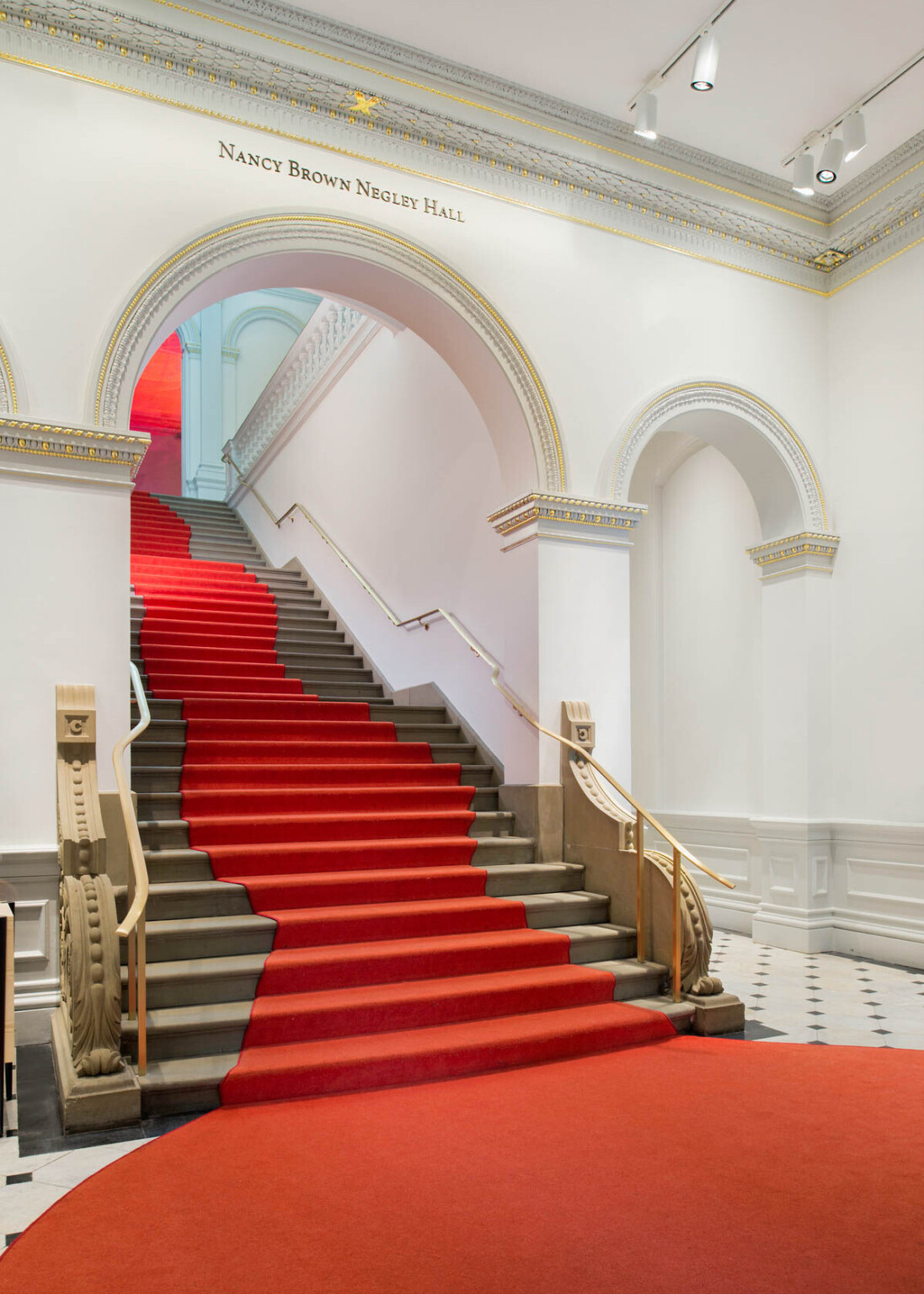Grand staircase with curved red carpet that widens at base of stairs. Stairs pass through a white arch with gold molding