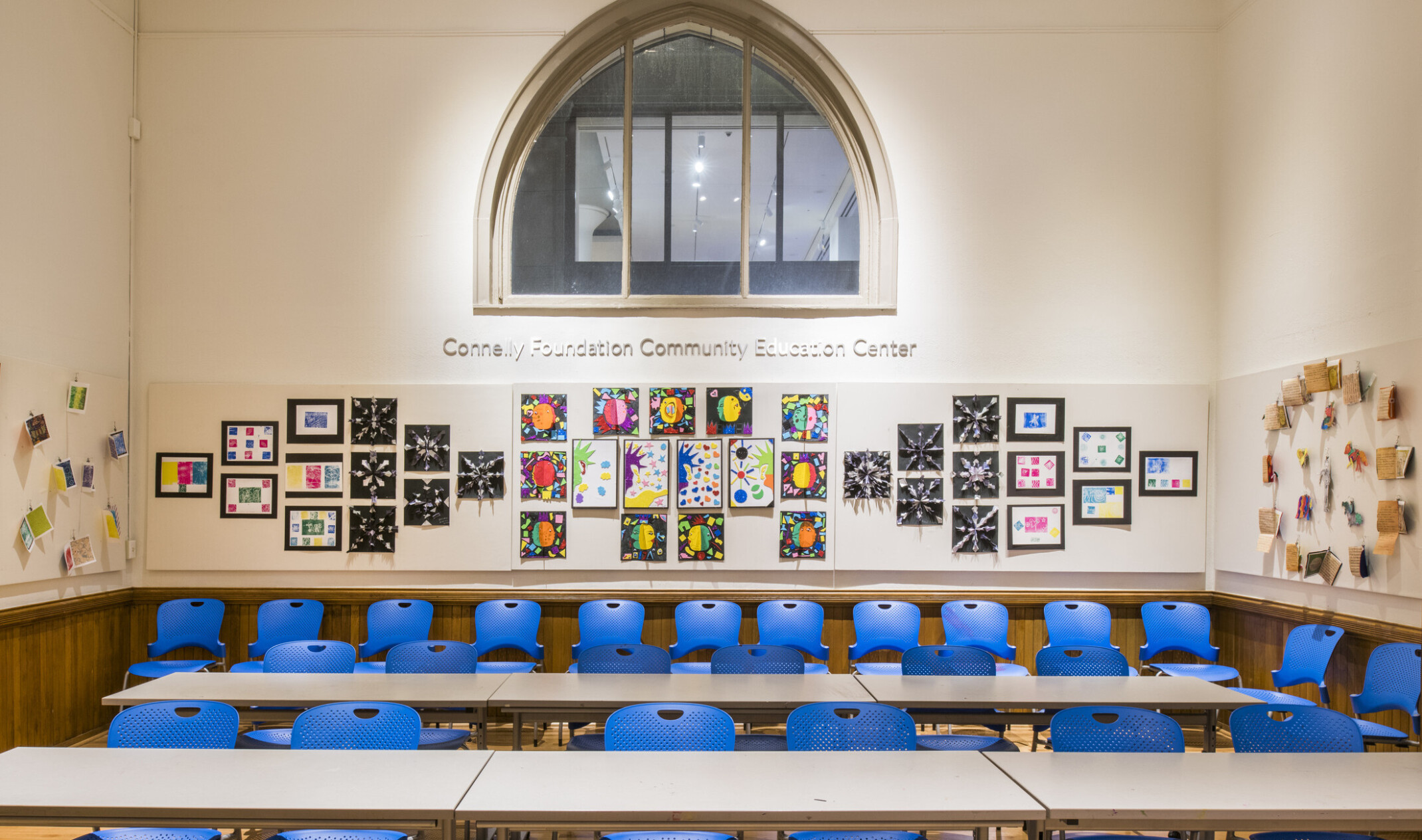 White room with pointed arch window at the center of the back wall. Tables in 2 lines with blue chairs fill the room