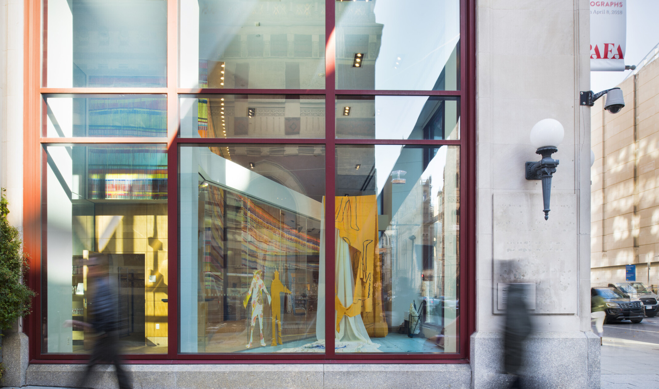 Double height windows with red trim seen from the street next to a light with metal sconce. An art gallery can be seen inside