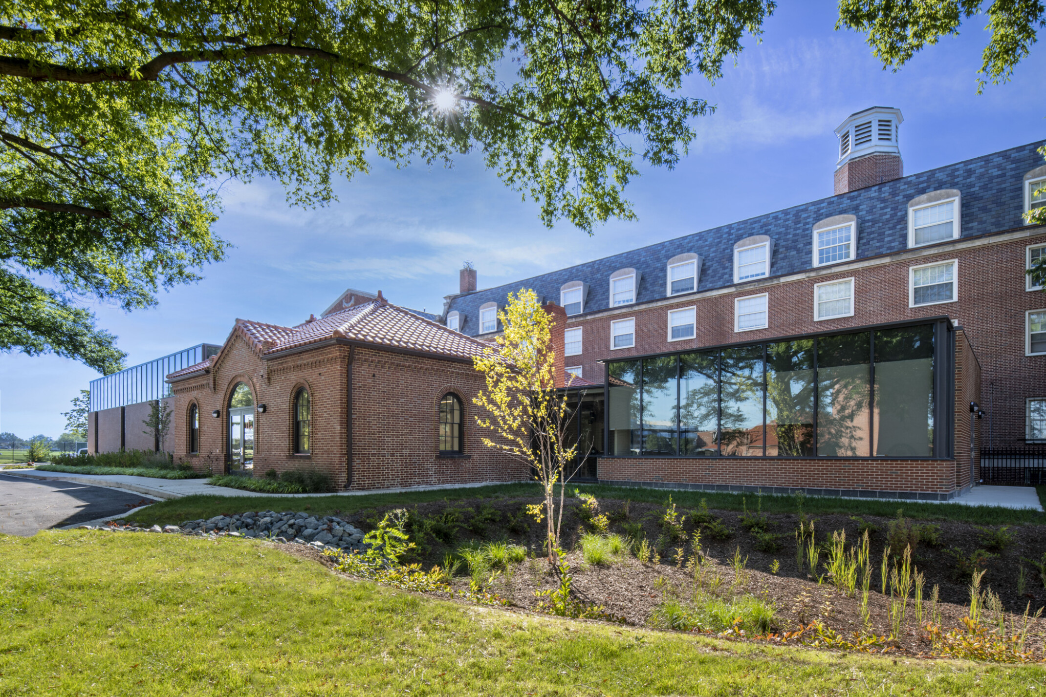 Exterior twilight view of the DDOT Materials Testing Lab showing red brick walls, double-door entrance, a glazed connecting corridor between the historic structure and new labs, and well-maintained native landscaping around the building.