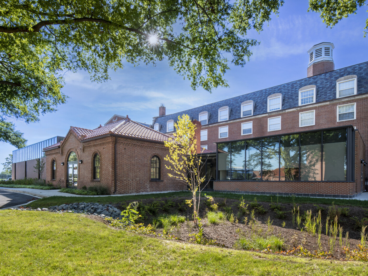 Exterior twilight view of the DDOT Materials Testing Lab showing red brick walls, double-door entrance, a glazed connecting corridor between the historic structure and new labs, and well-maintained native landscaping around the building.