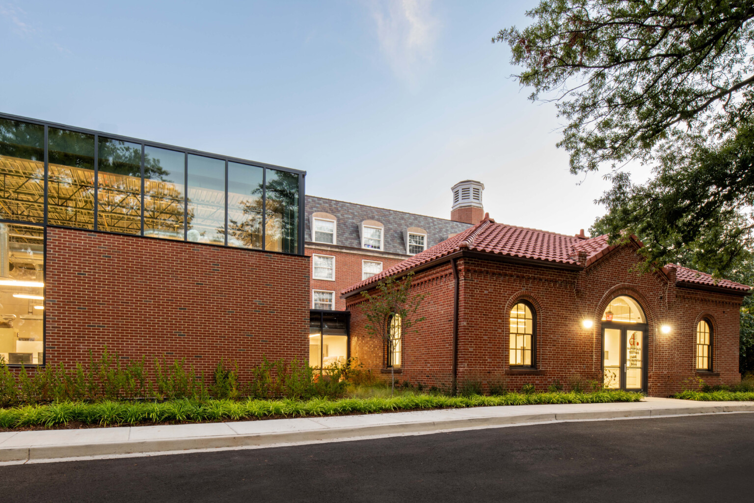 Exterior twilight view of the DDOT Materials Testing Lab showing red brick walls, double-door entrance, a glazed connecting corridor between the historic structure and new labs, and well-maintained native landscaping around the building.