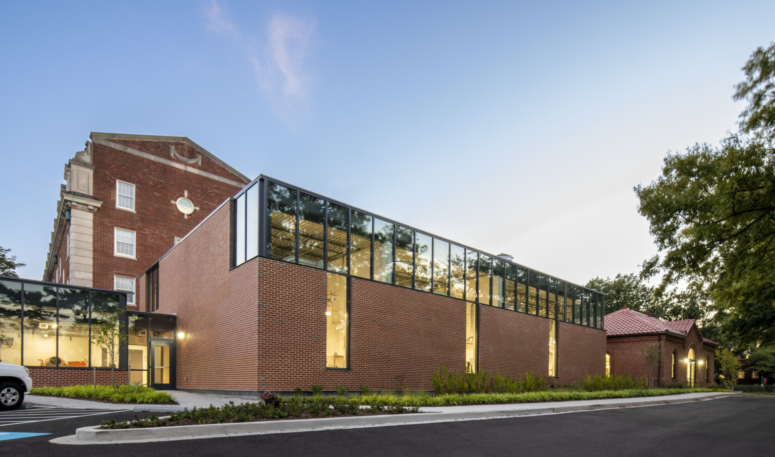 Exterior twilight view of the DDOT Materials Testing Lab showing red brick walls, double-door entrance, a glazed connecting corridor between the historic structure and new labs, and well-maintained native landscaping around the building.
