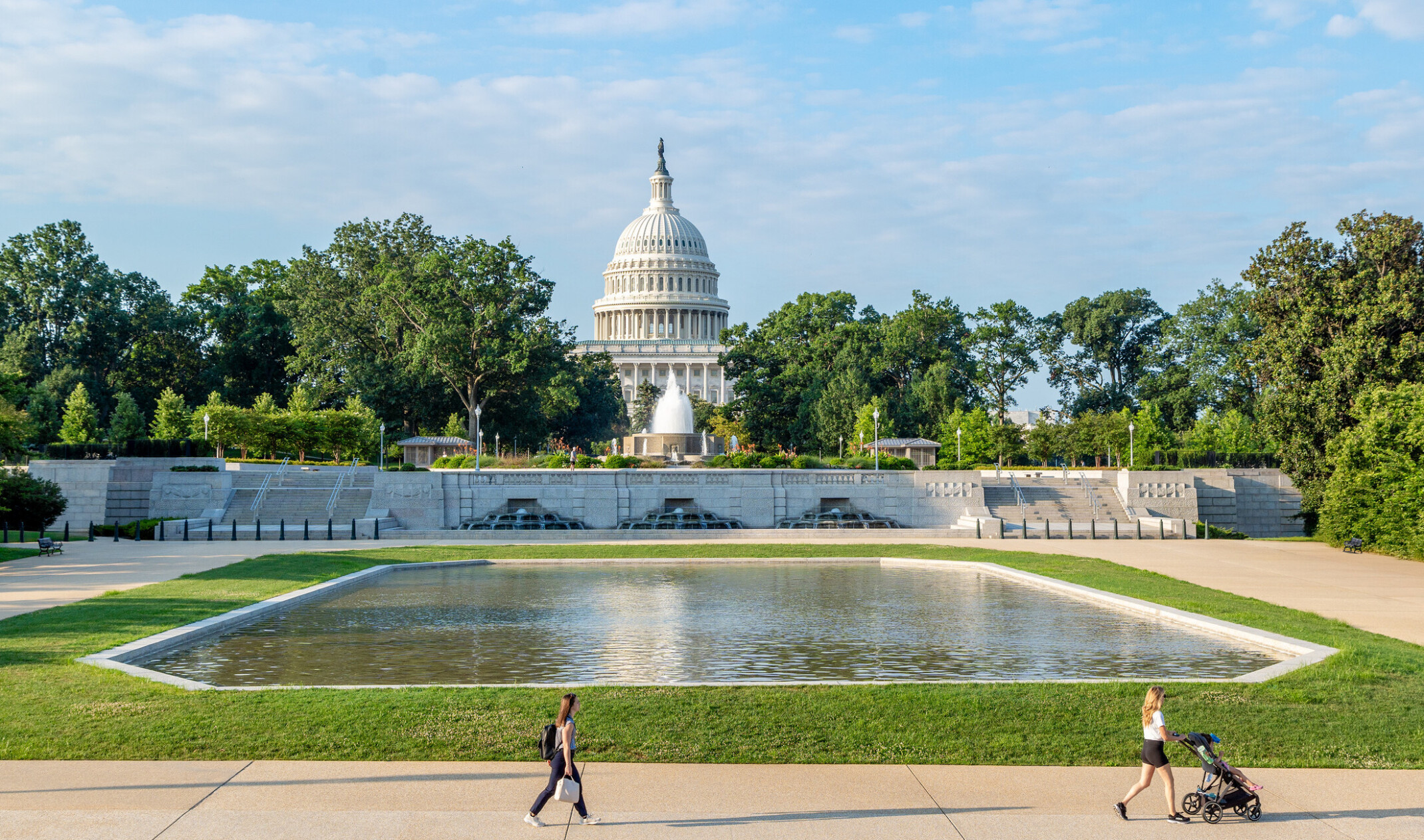 A wide, street-level shot of the reflection pool with patrons walking about in the foreground, featuring the Capitol dome and fountains in the background