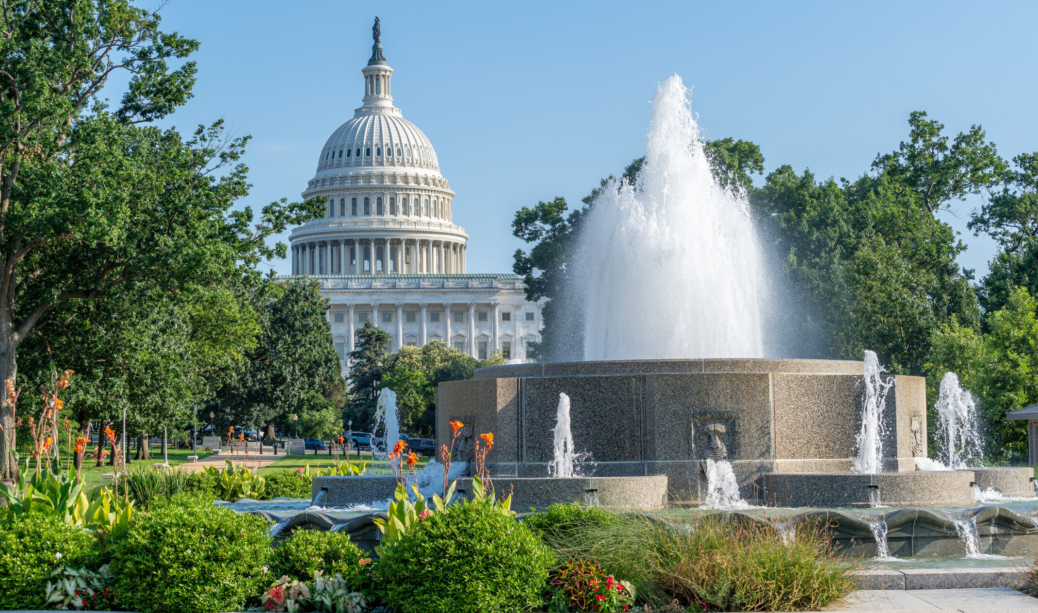 A marble water fountain with water cascading sits before the Capitol dome surrounded by lush trees and landscaping.