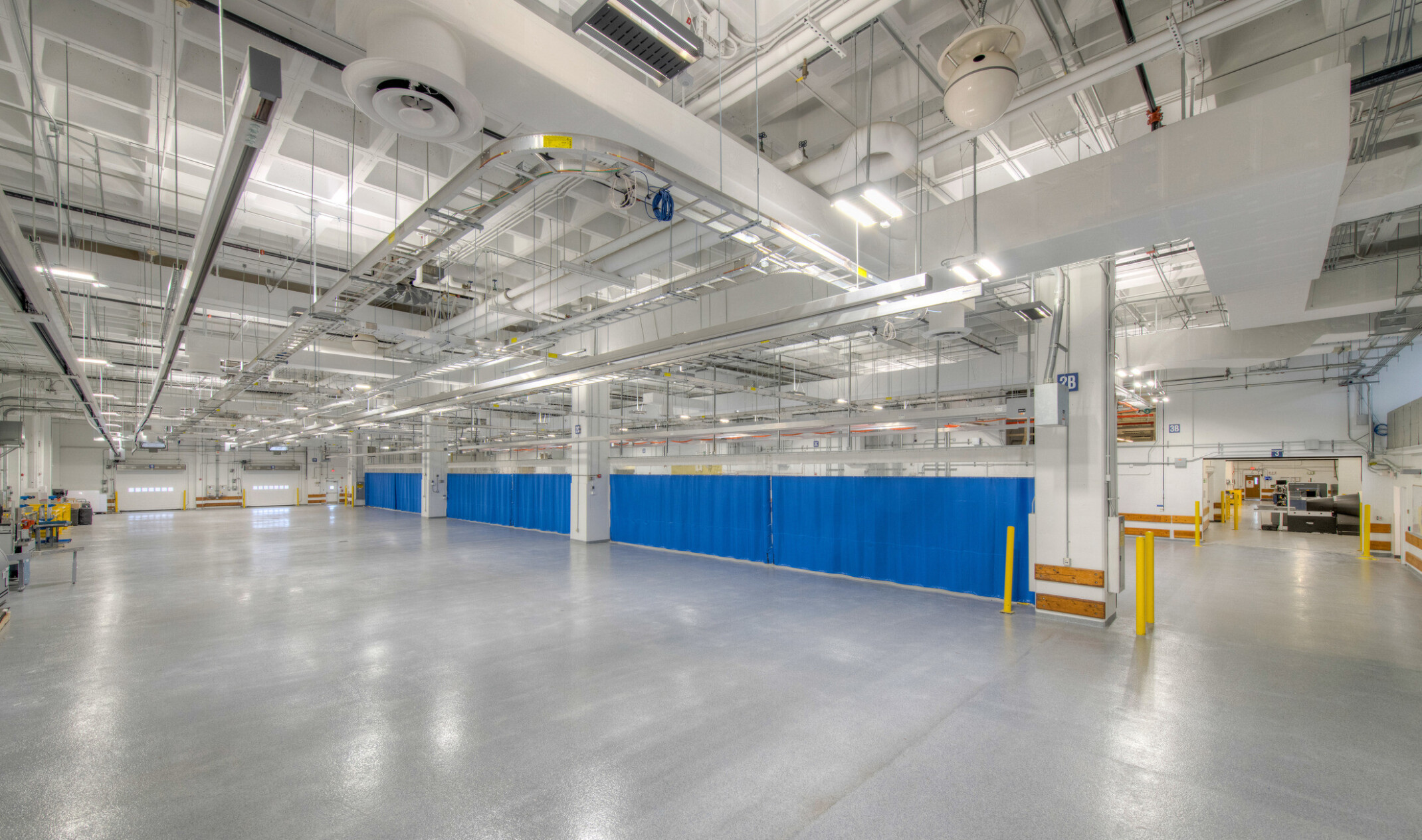 Interior of a windowless testing facility with polished concrete floors, exposed ceiling pipes, bright safety-coded elements, and overhead lighting.