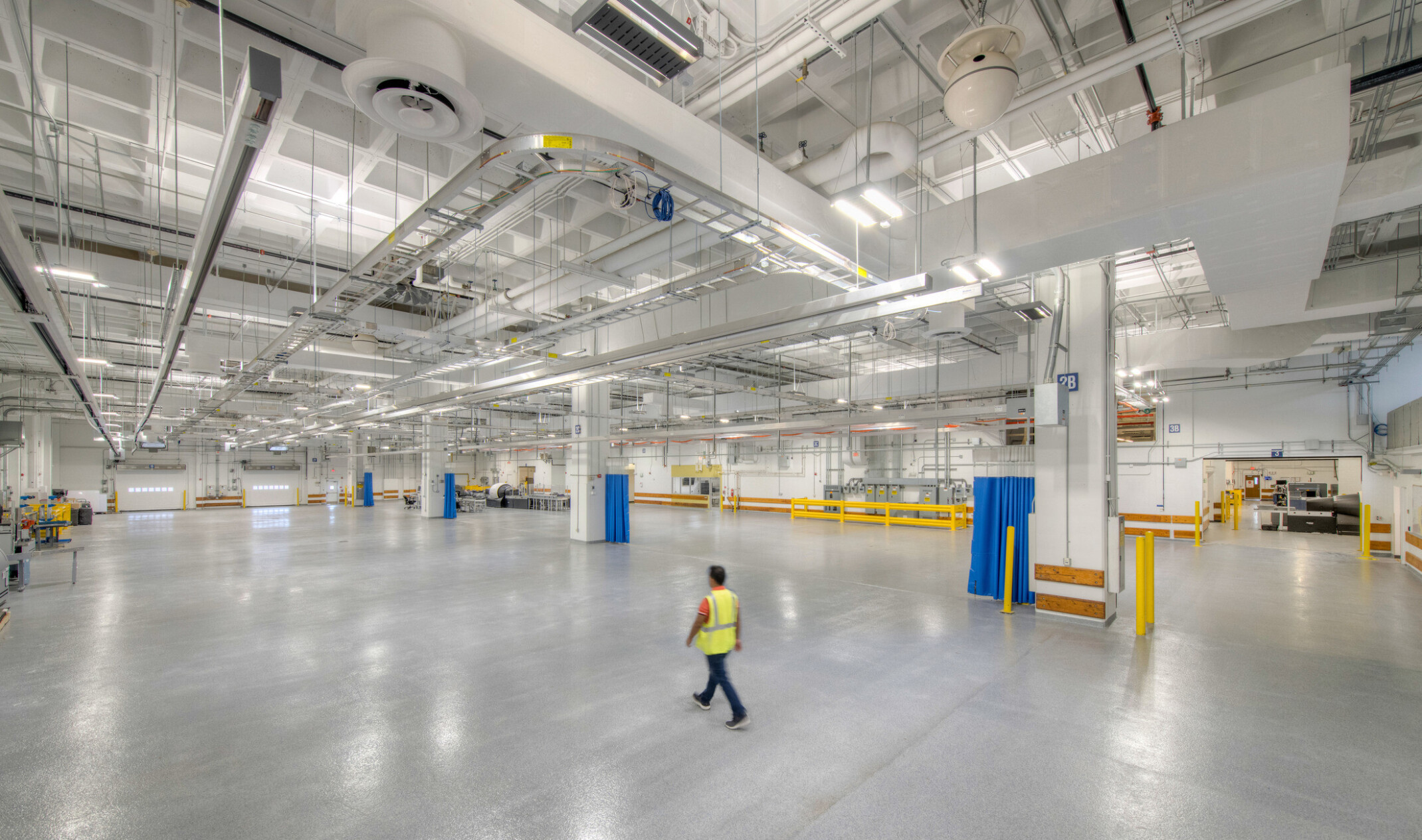 Interior of a windowless testing facility with polished concrete floors, exposed ceiling pipes, bright safety-coded elements, and overhead lighting.
