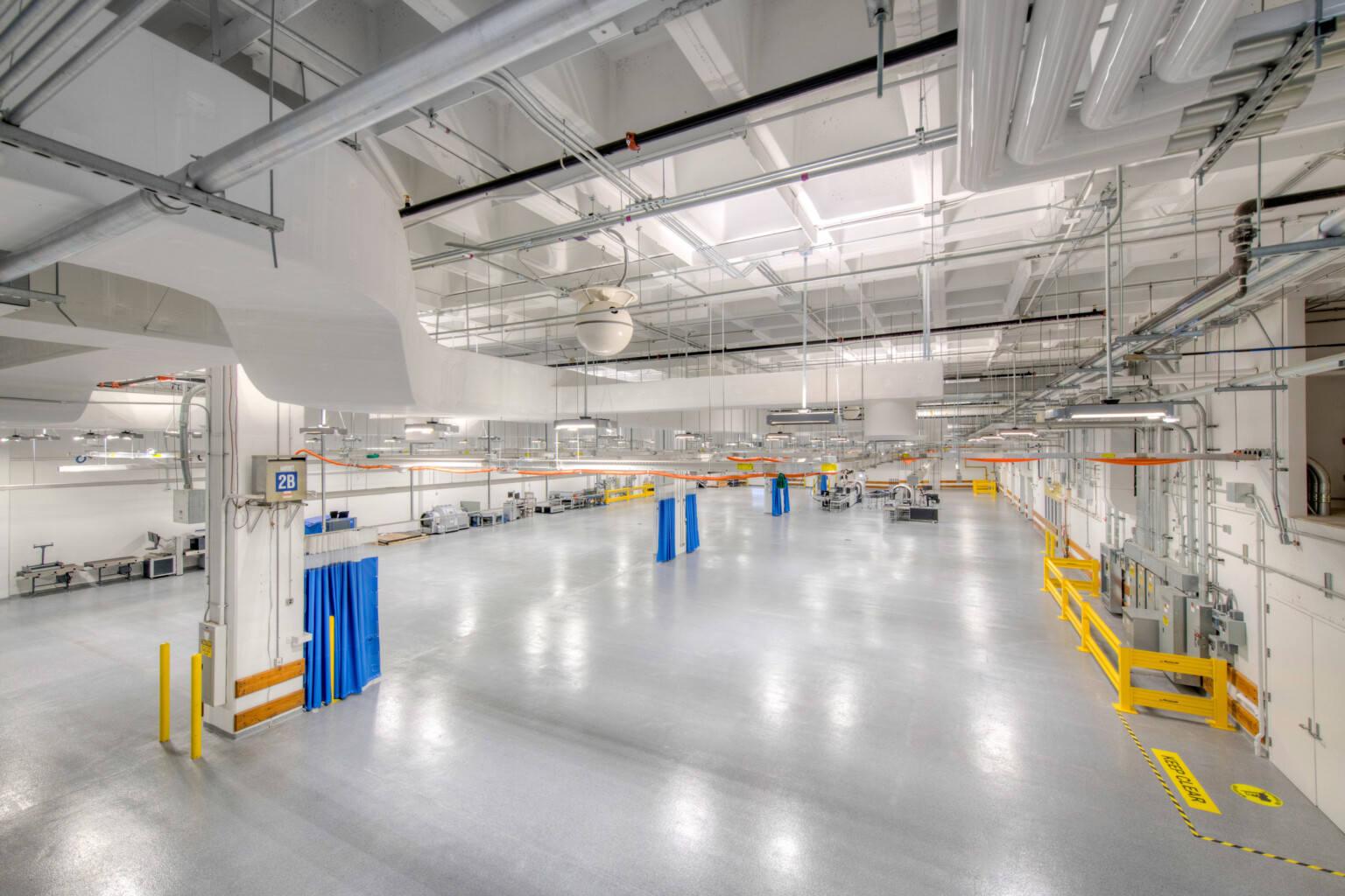 Interior of a windowless testing facility with polished concrete floors, exposed ceiling pipes, bright safety-coded elements, and overhead lighting.