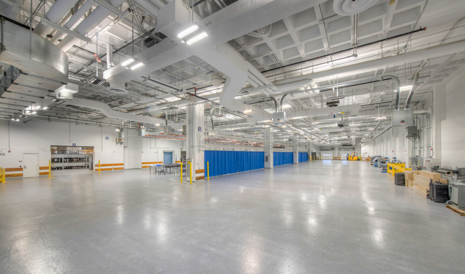 Interior of a windowless testing facility with polished concrete floors, exposed ceiling pipes, bright safety-coded elements, and overhead lighting. Interior of a windowless testing facility with polished concrete floors, exposed ceiling pipes, bright safety-coded elements, and overhead lighting.