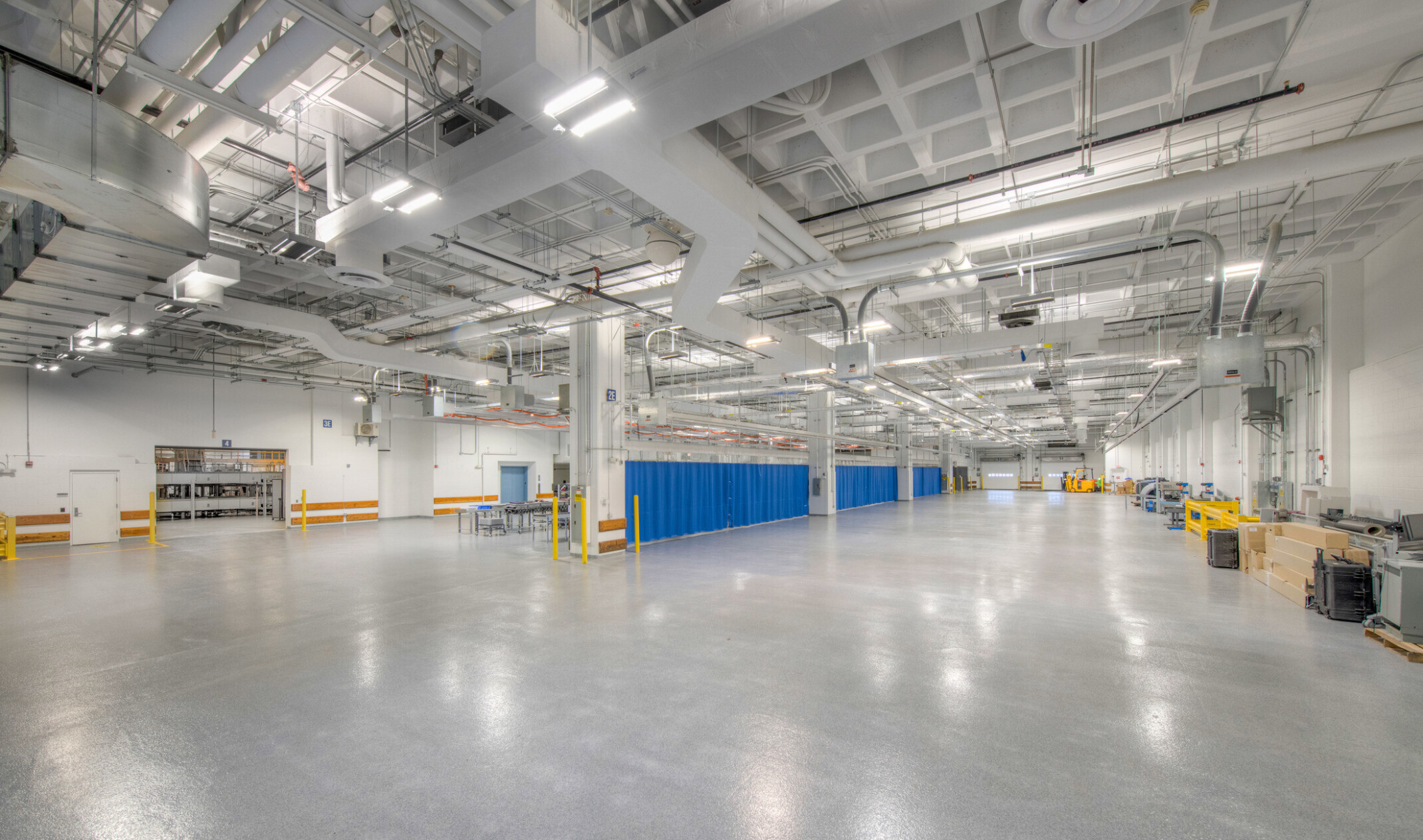 Interior of a windowless testing facility with polished concrete floors, exposed ceiling pipes, bright safety-coded elements, and overhead lighting.
