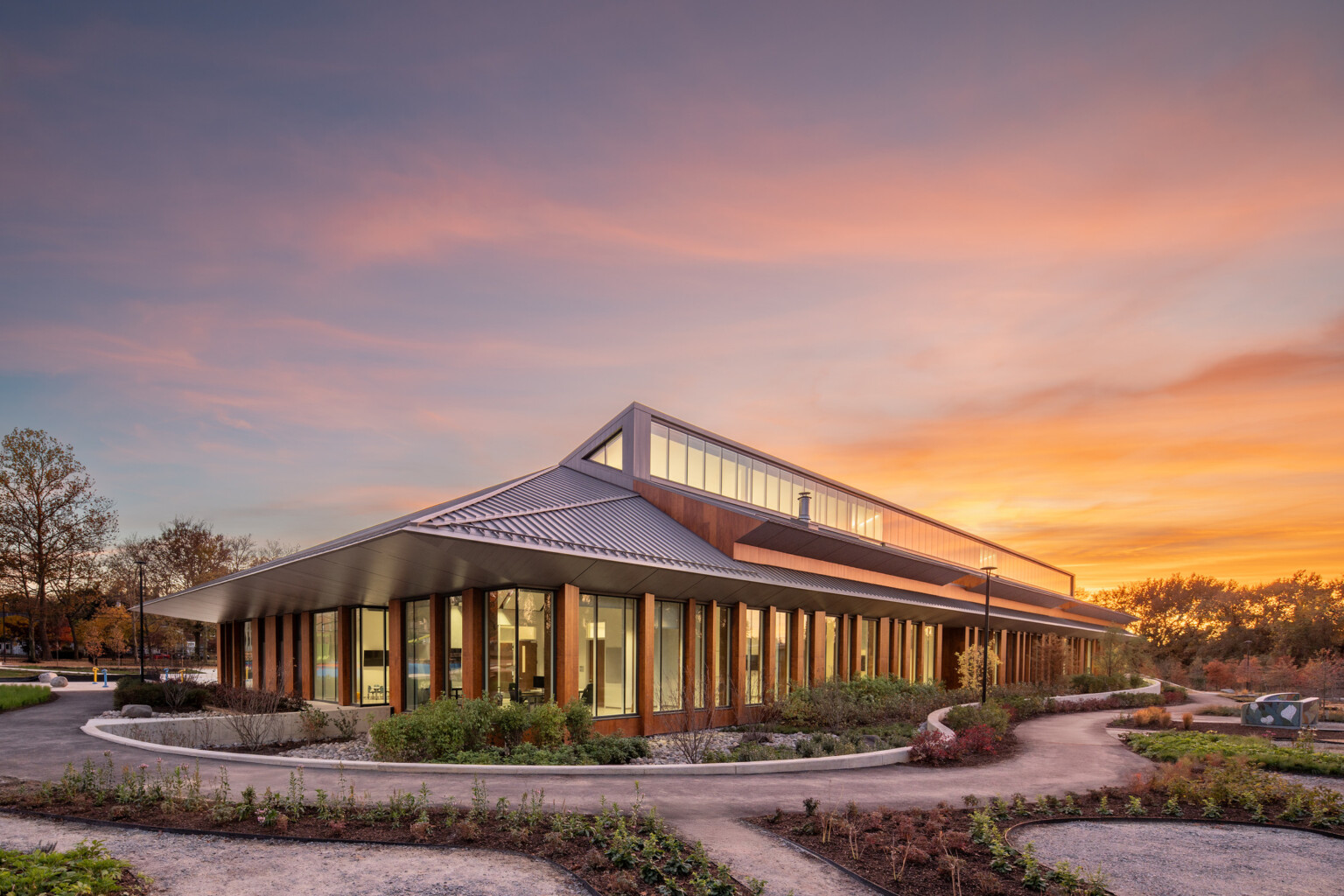 Modern, angular building with a slanted metal roof and large glass windows surrounded by lush landscaping at sunset