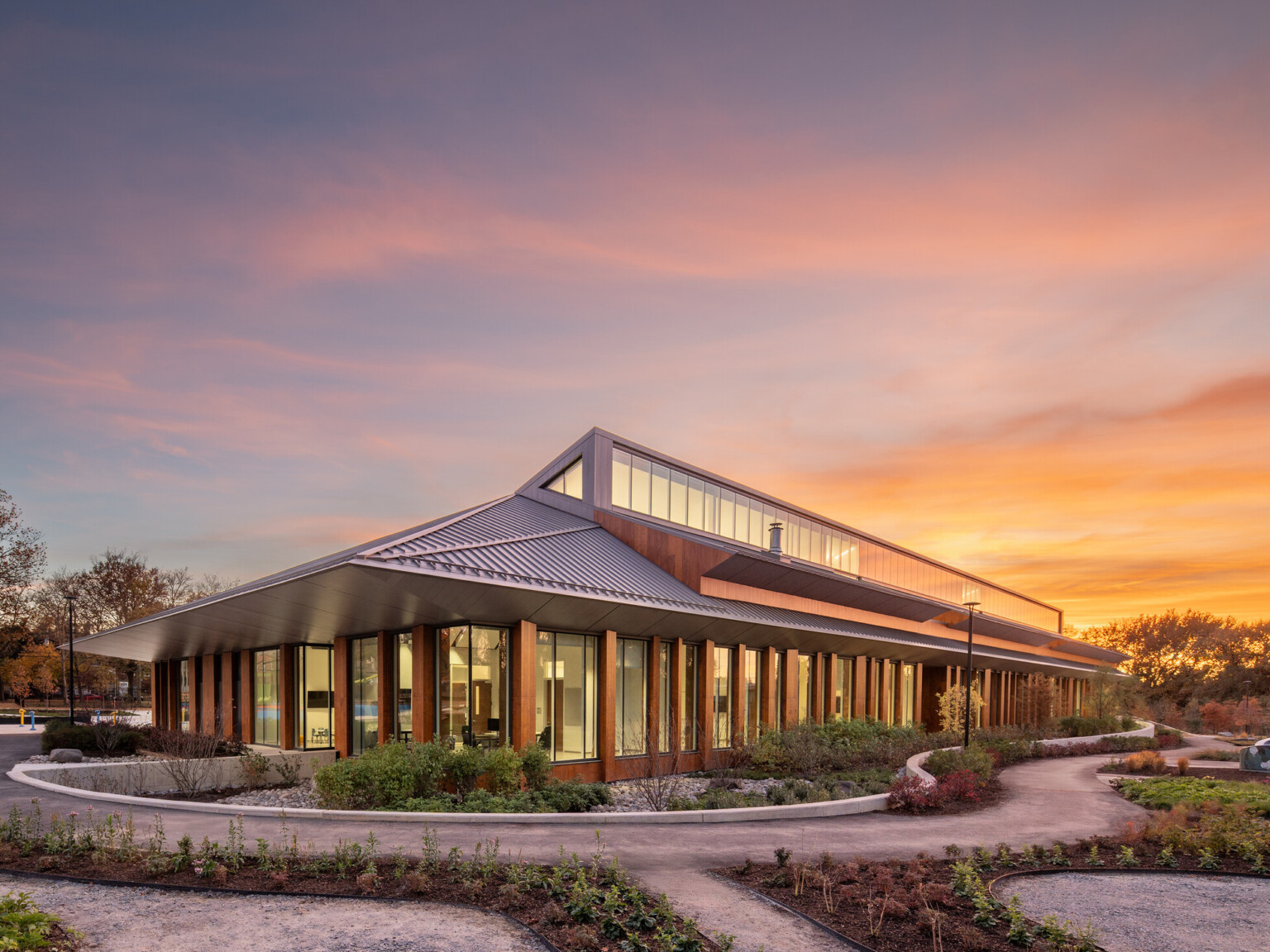 Modern, angular building with a slanted metal roof and large glass windows surrounded by lush landscaping at sunset