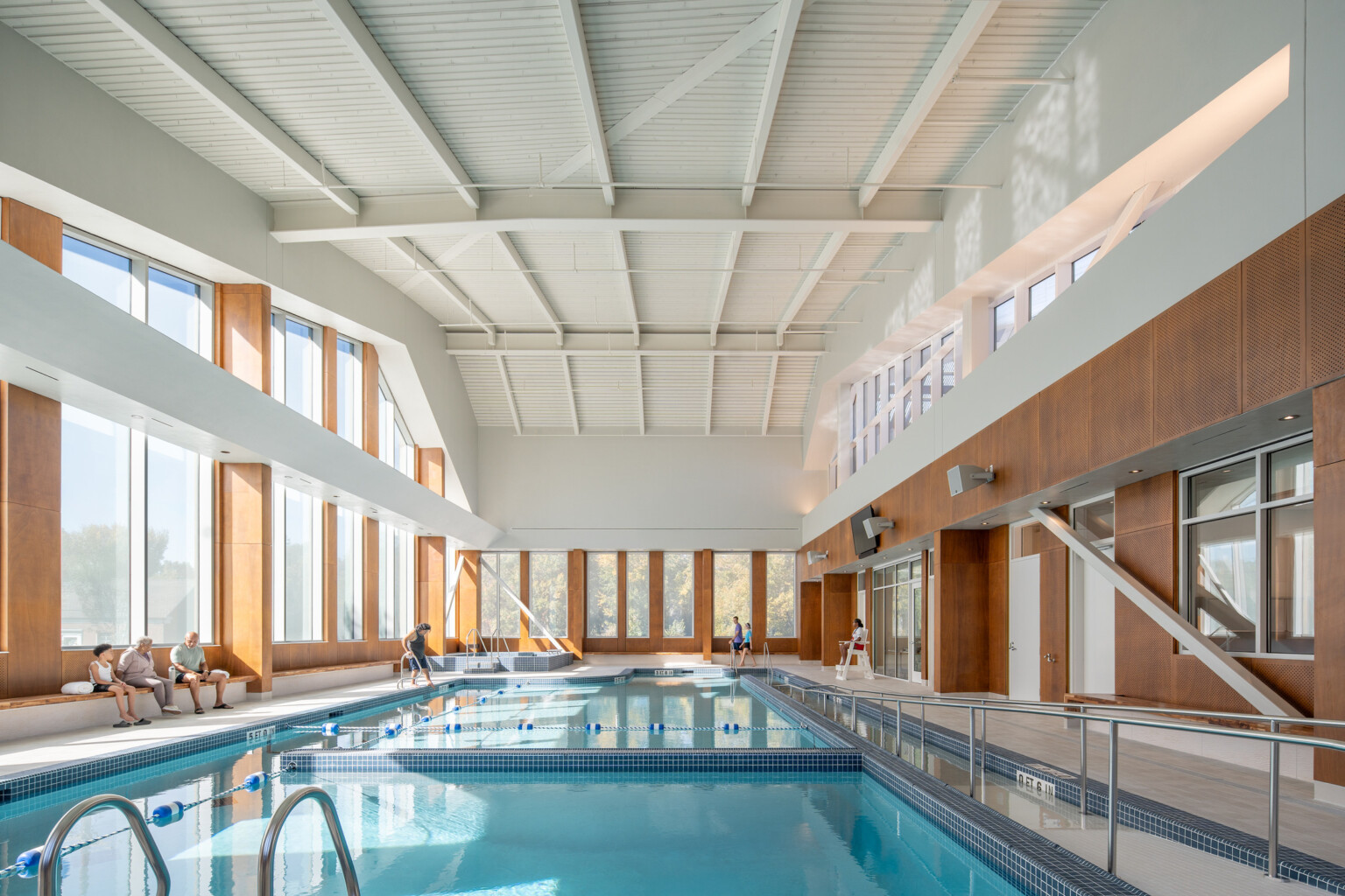 Indoor swimming pool with large windows and wooden accents, featuring several people sitting and standing nearby.
