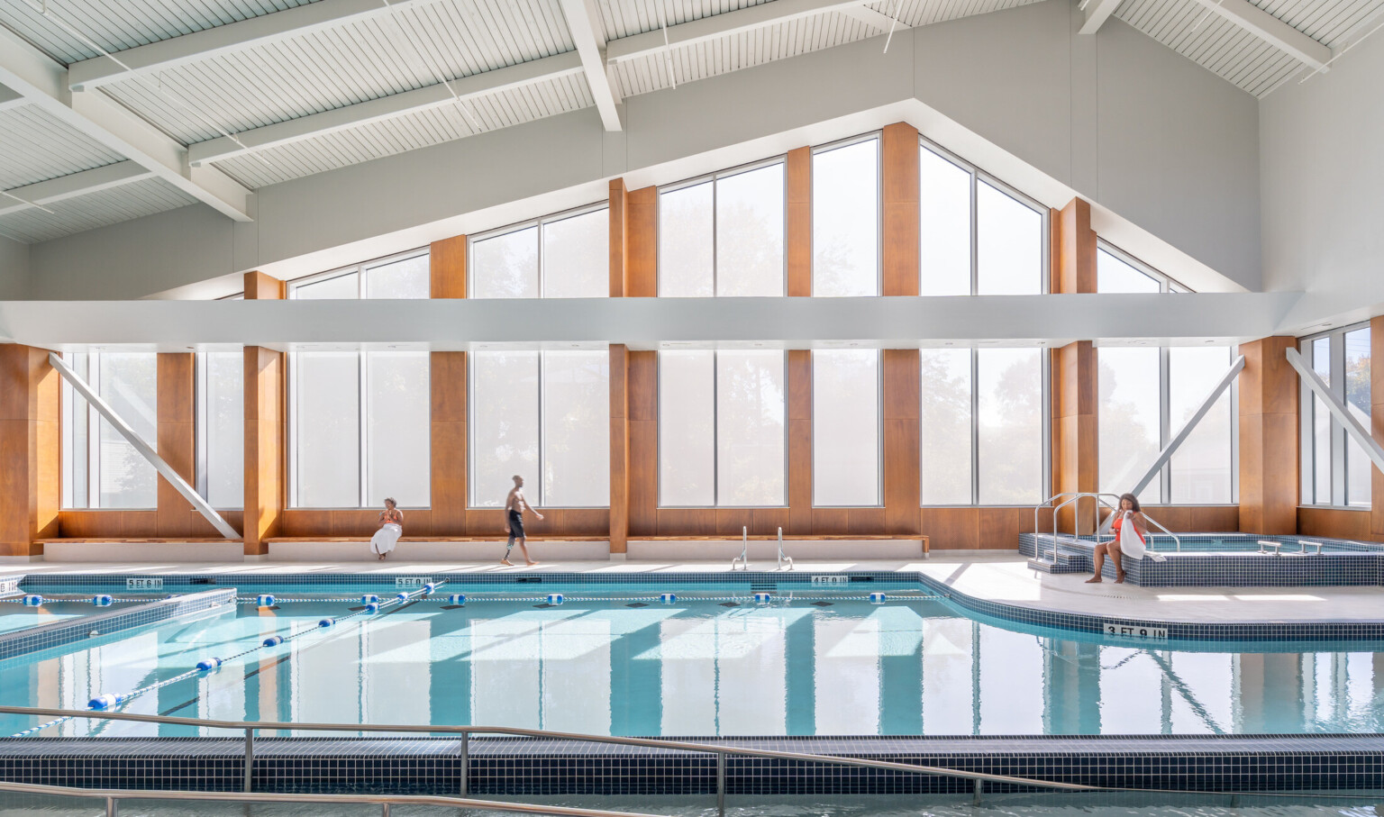 Indoor swimming pool with large windows and wooden accents, featuring several people sitting and standing nearby.