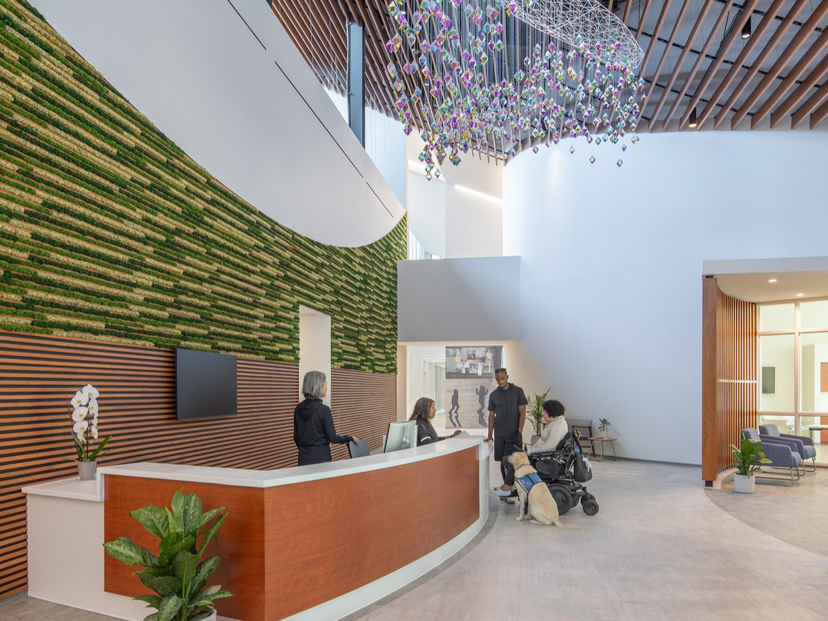 tall lobby with a large reception desk on the left, backed by a wall featuring grasses set between wooden slats. An iridescent chandelier hangs from the high ceiling in front of the desk. Two staff members are behind the desk, with one assisting two visitors, one standing and one in a wheelchair with a service dog.