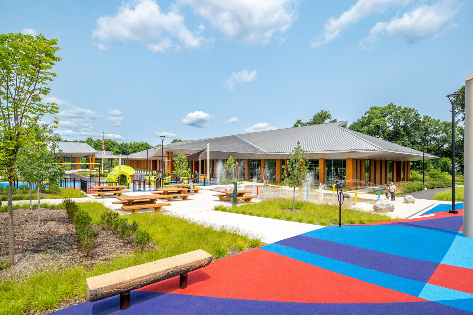 splashpad in front of a recreation center on a warm sunny day; children and grandparents playing with water features; rubber-surface playground with accessible playground equipment; park benches that are shaped to resemble split logs; fenced toddler play area under shade structure