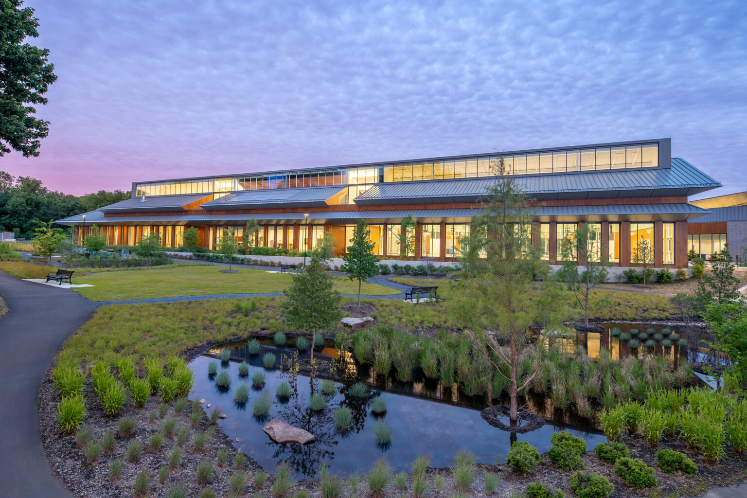 Outdoor view of a contemporary recreation center made of wood, steel, and large windows, surrounded by native plants and manicured landscaping under the light of dusk with scattered clouds reaching across the sky.