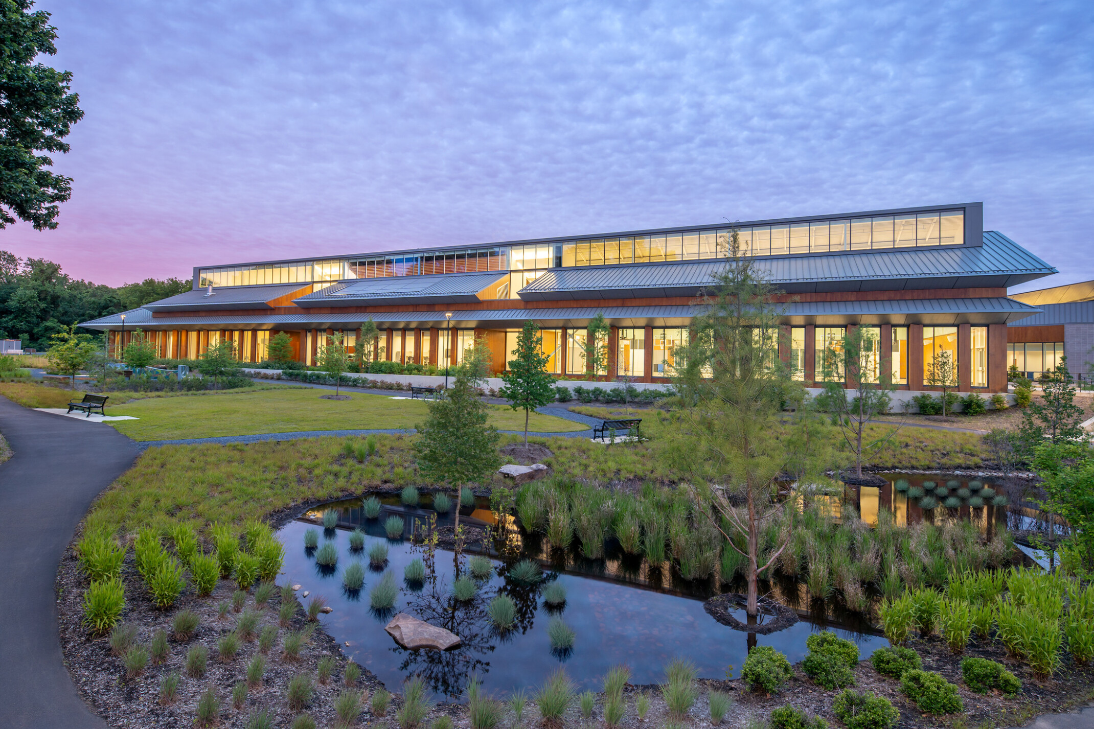 Outdoor view of a contemporary recreation center made of wood, steel, and large windows, surrounded by native plants and manicured landscaping under the light of dusk with scattered clouds reaching across the sky.