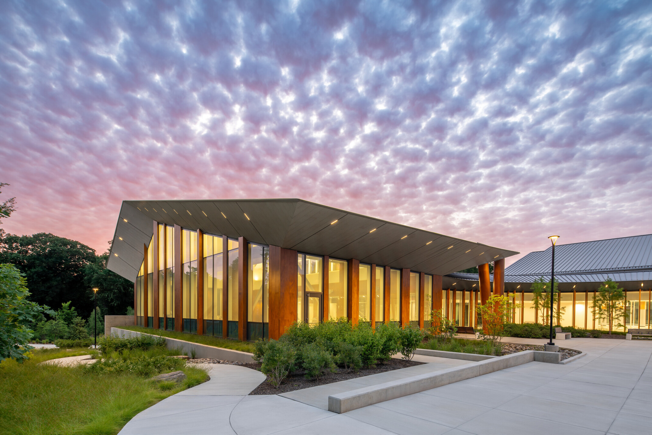 Exterior of rec center at dusk, with windows glowing under a purple sunset sky. White sidewalks meander around the building, through areas with native plants and low bushes