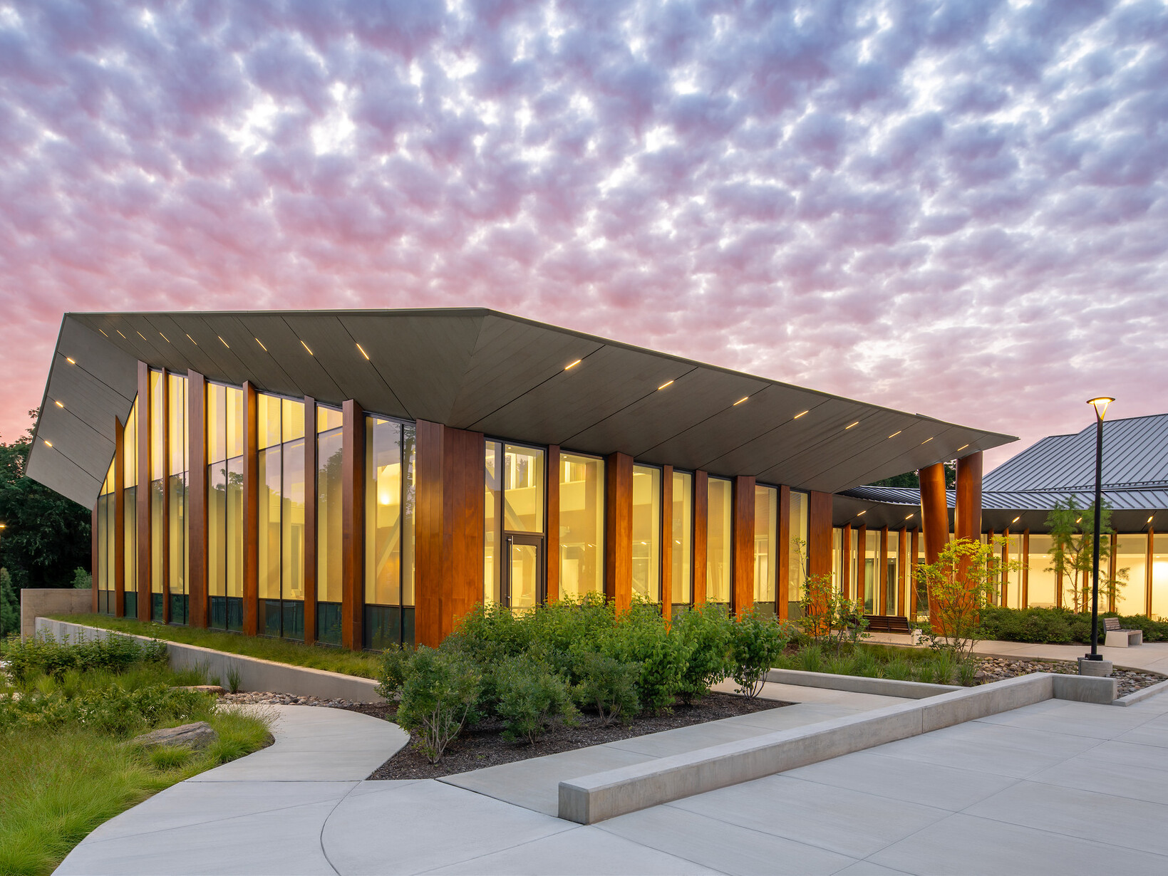 Exterior of rec center at dusk, with windows glowing under a purple sunset sky. White sidewalks meander around the building, through areas with native plants and low bushes
