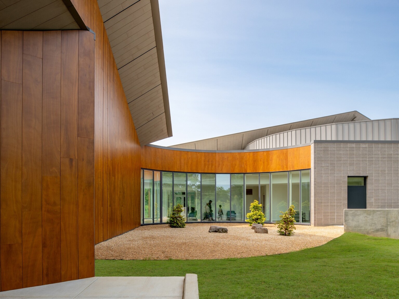 Semicircular exterior courtyard wrapped by the wood and window-clad exterior of the building. The courtyard surface features gravel, small boulders, and low evergreen trees, bounded by lush, short grass.