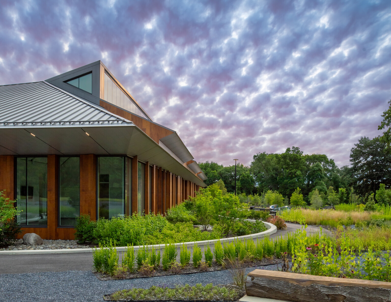 One-story rec center building with heavy timber-like columns, large windows, and metal roof; a clerestory runs the length of the building; the building is surrounded by gravel paths winding between flowering native gardens; metal benches sit along the paths