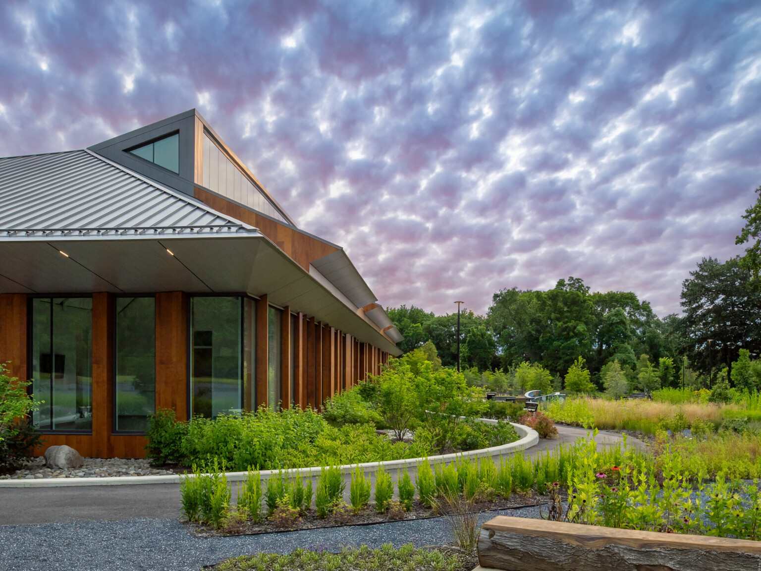 One-story rec center building with heavy timber-like columns, large windows, and metal roof; a clerestory runs the length of the building; the building is surrounded by gravel paths winding between flowering native gardens; metal benches sit along the paths