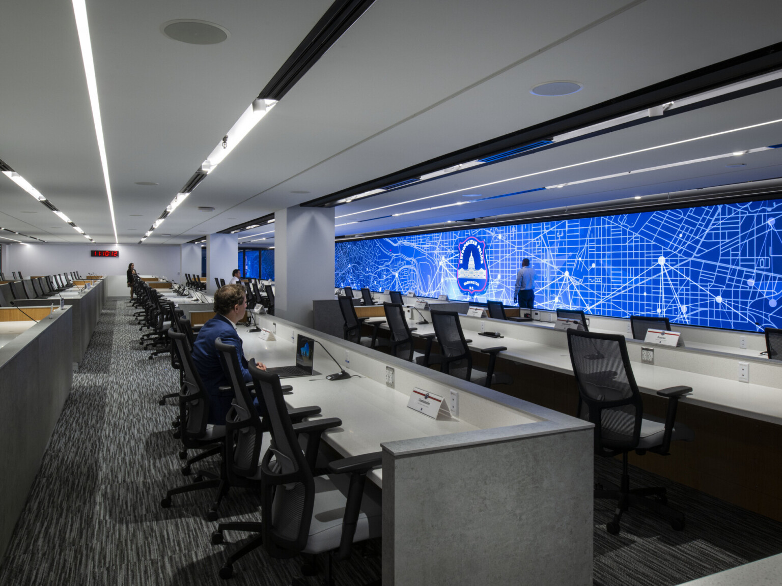 A modern police control room with multiple rows of workstations and a large digital map display on the wall