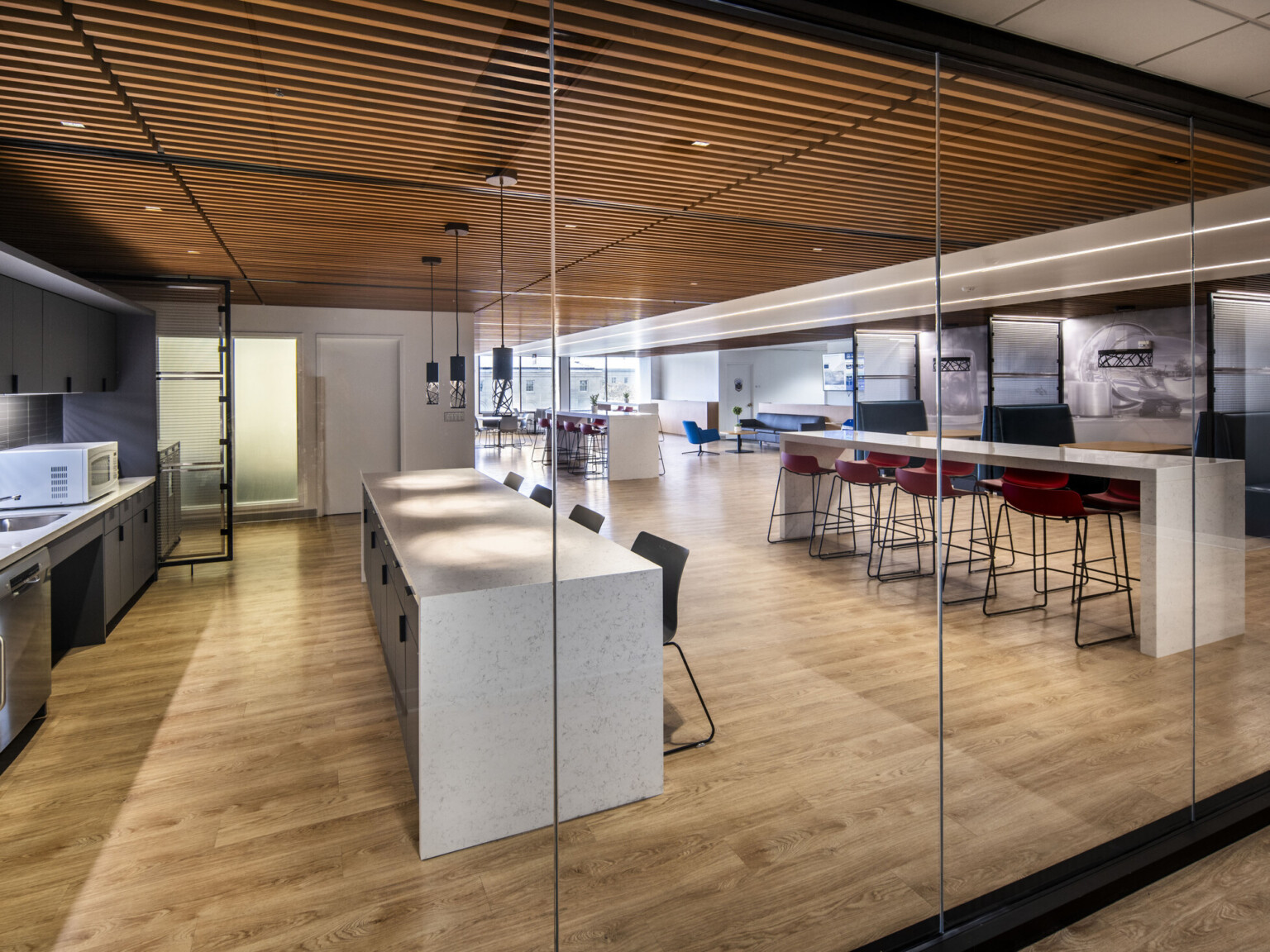 Modern office kitchen with dark cabinetry, white countertops, light wood floors, and adjacent seating area with red and black chairs