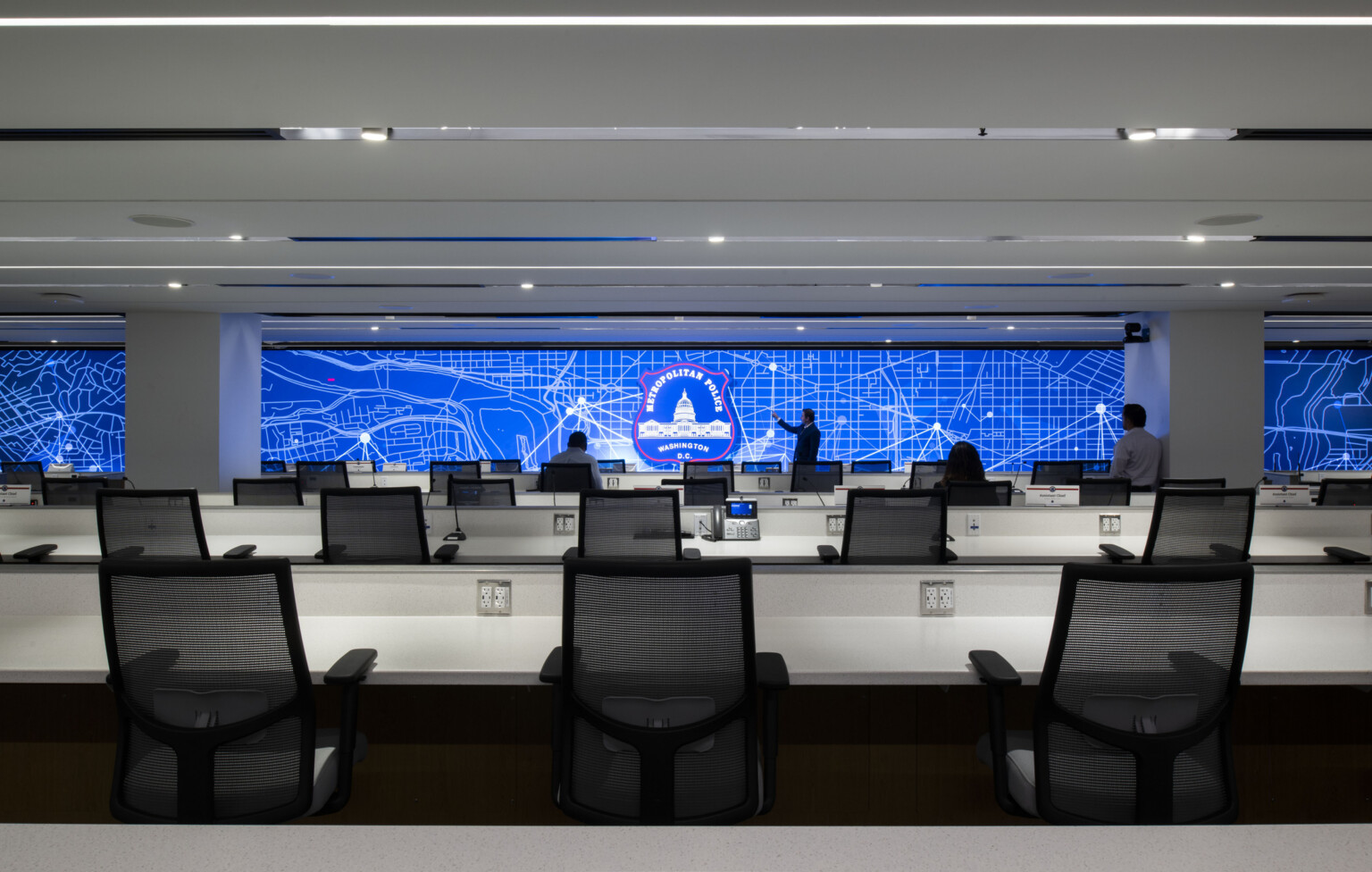 Modern police command center with rows of desks lined with chairs and a large digital map display on the back wall