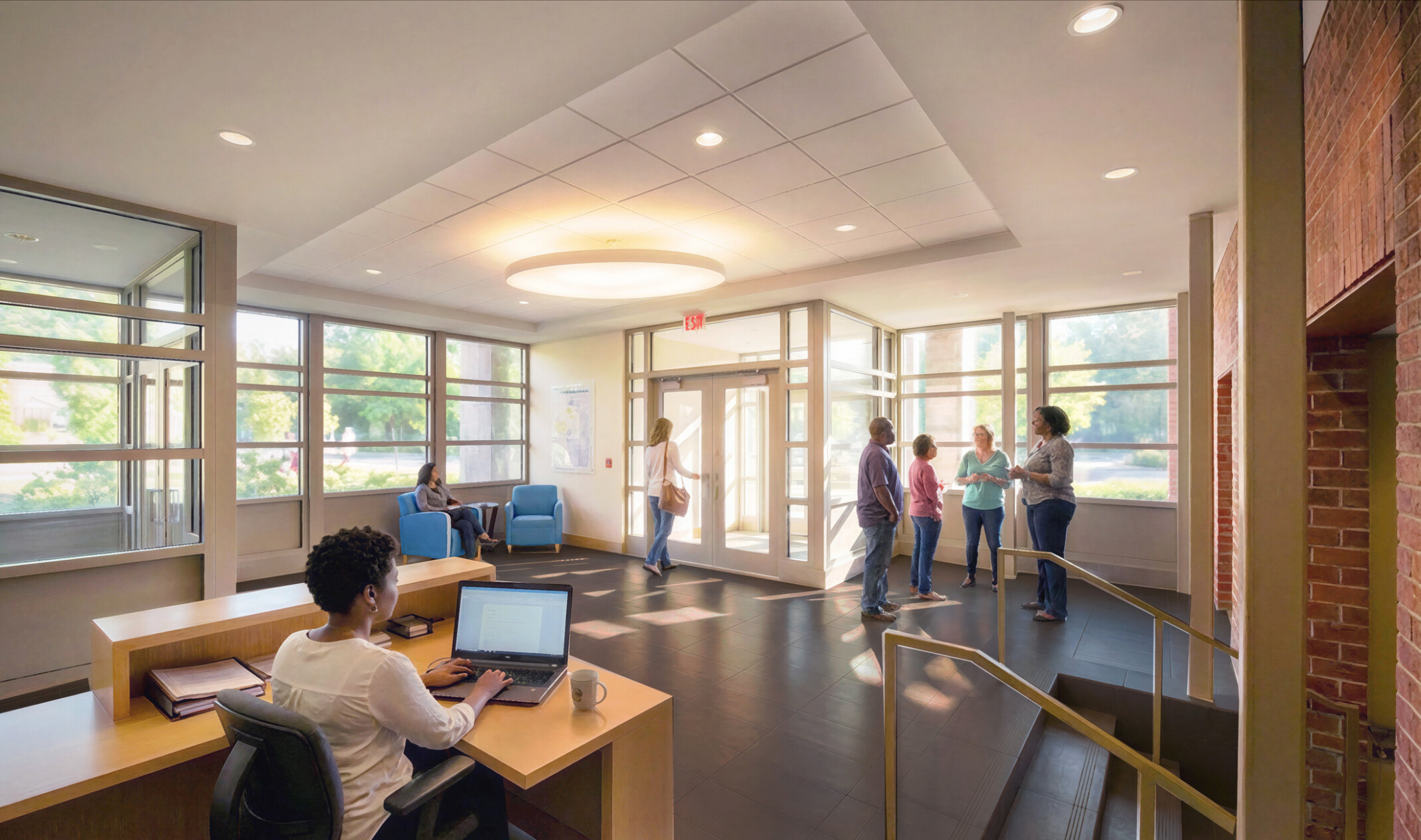 A lobby entrance with several windows allowing natural light, featuring a receptionist at a computer in front of a desk, people mingling in the lobby area, grey floors, recessed lighting, and natural brick walls from an original 1900s building.