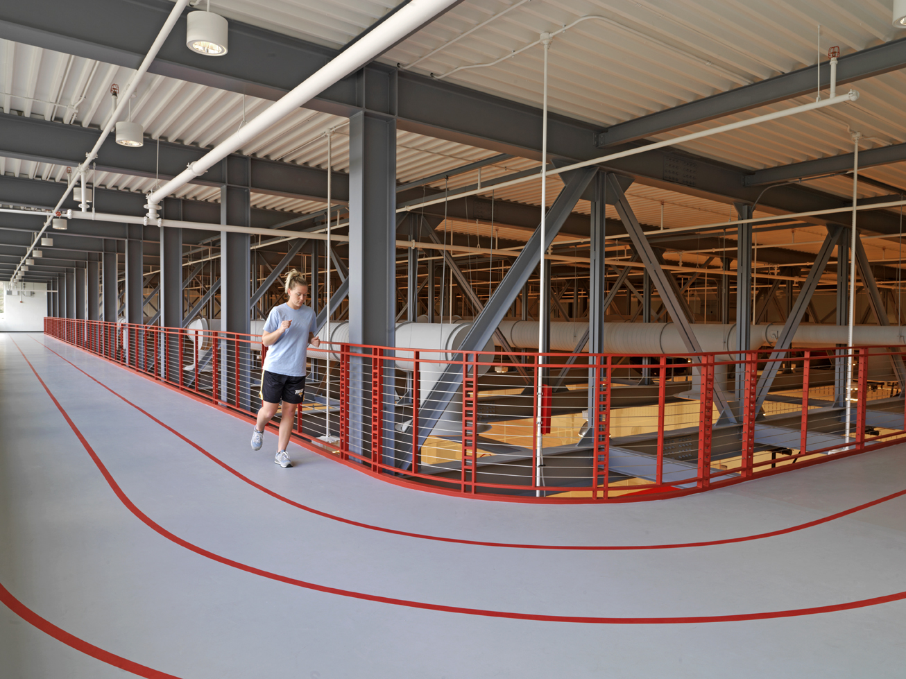 An individual running on an indoor track with grey floors with lanes divided by red lines in front of red railings