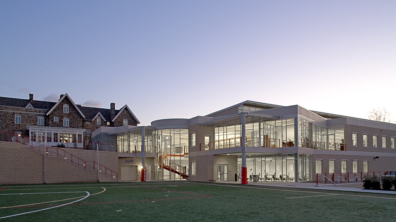 Athletic building filled with well lit windows overlooking a green grass playing field with a rock multistory building in the backdrop