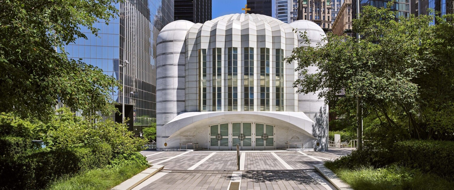 Daytime shot of the St. Nicholas Greek Orthodox Church New York. White building surrounded by trees and tall glass buildings
