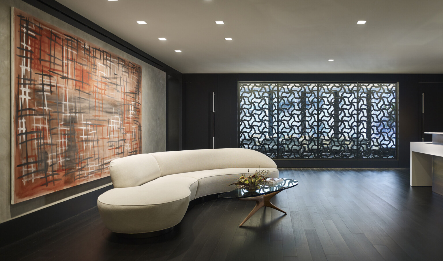 Curved white couch with glass coffee table in wood room with abstract art behind. Back wall has organically patterned panels