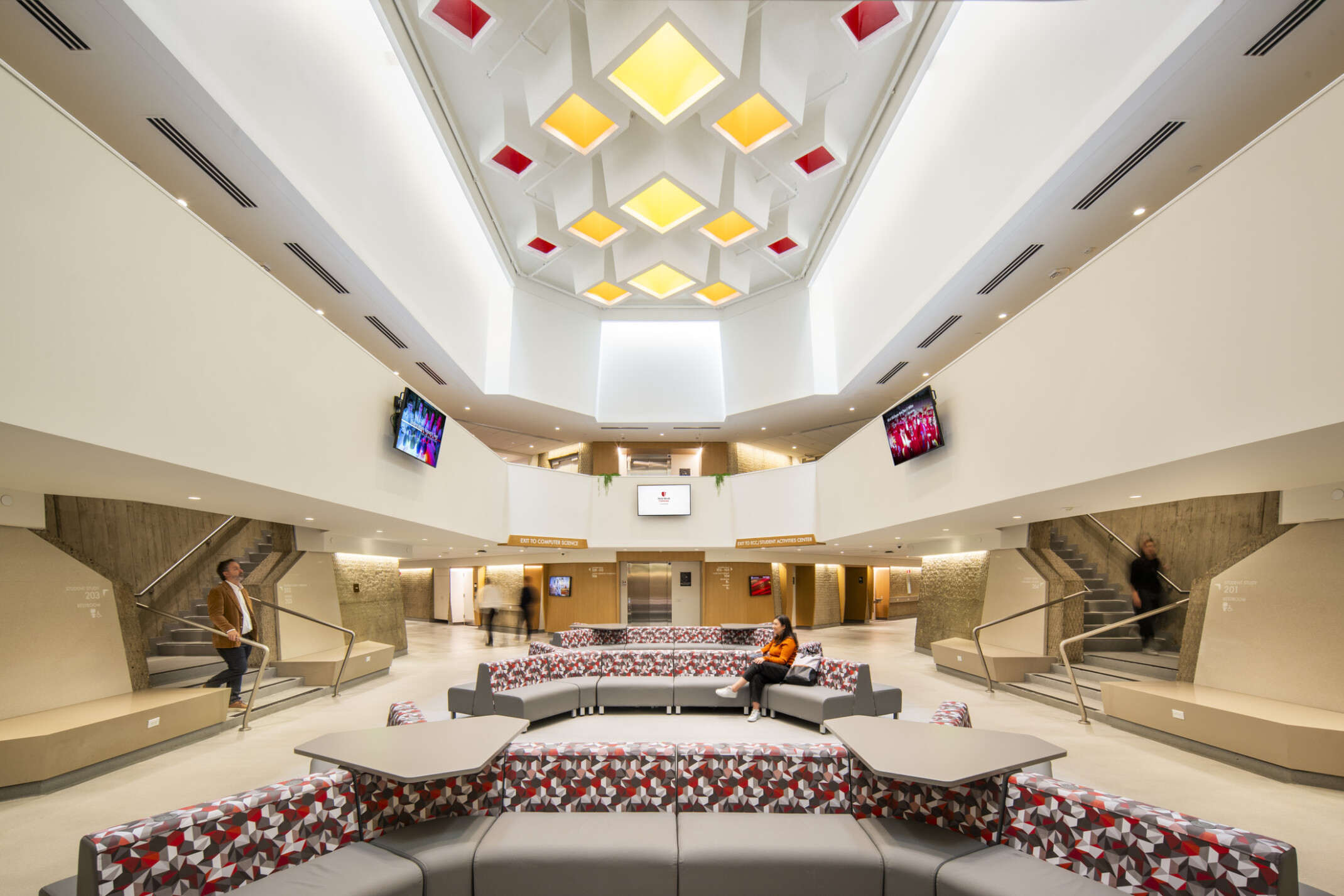 A spacious, modern lobby with high ceilings and geometric recessed lighting panels; large circular seating arrangement with patterned cushions and attached tables; 2people seated on the couches;2 others walking near staircases; light-colored walls ;concrete accents; flat-screen monitors mounted on both sides displaying colorful graphics.