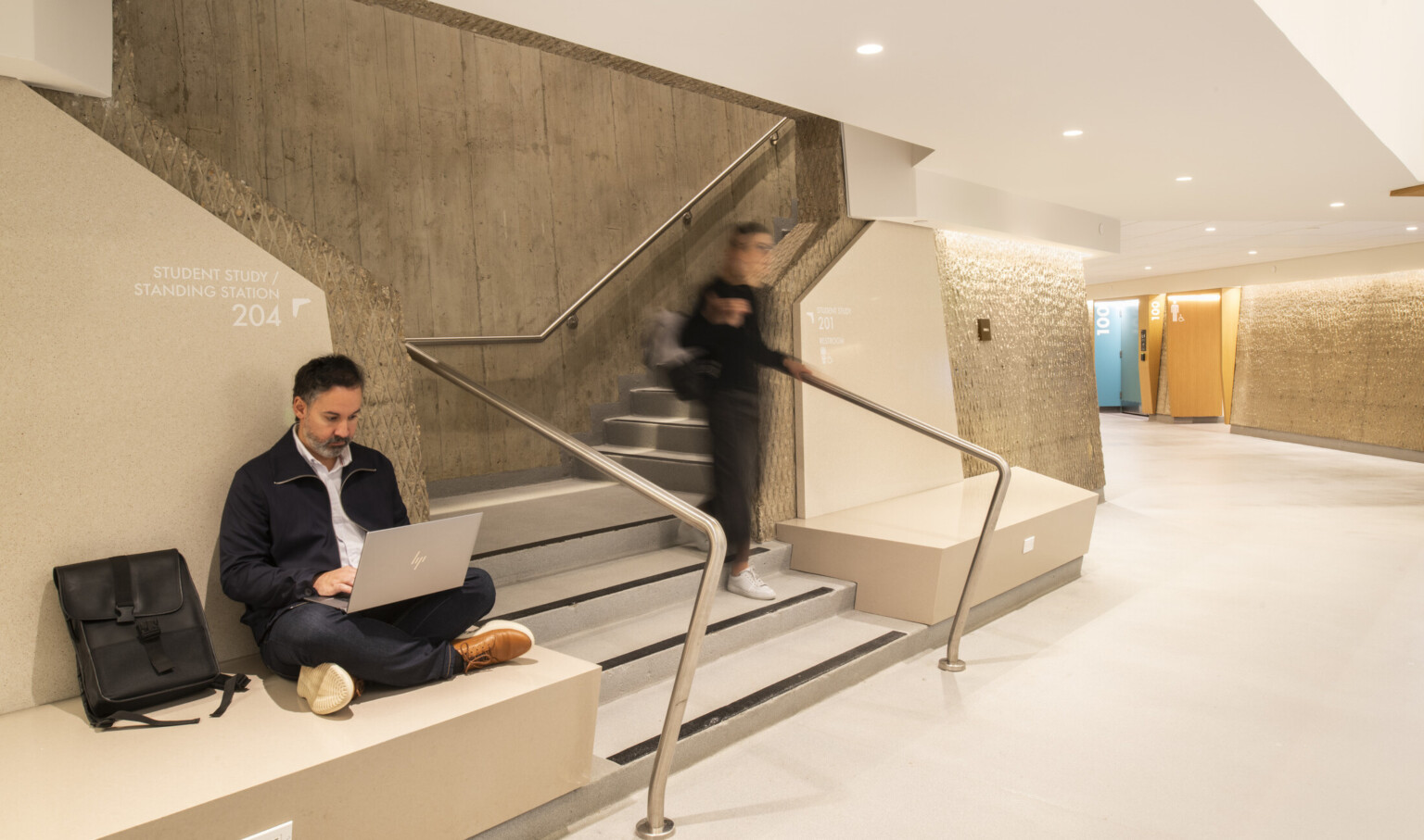 modern interior hallway with light-colored walls and flooring; person seated on a ledge near a staircase, working on a laptop with a black bag;; another person walking down the stairs, appearing blurred from motion; staircase with sleek metal handrail and concrete wall backdrop; hallway curves with brightly colored doors visible