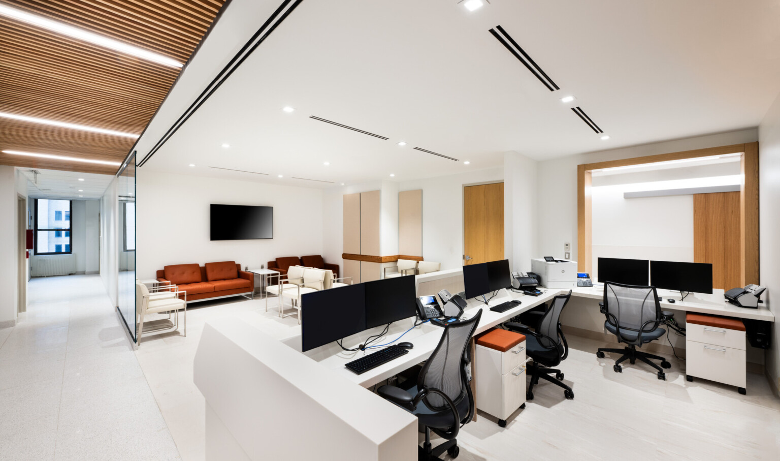 View of a medical office waiting room with a wooden doors with computers on a desk and off-white chairs in the lobby area