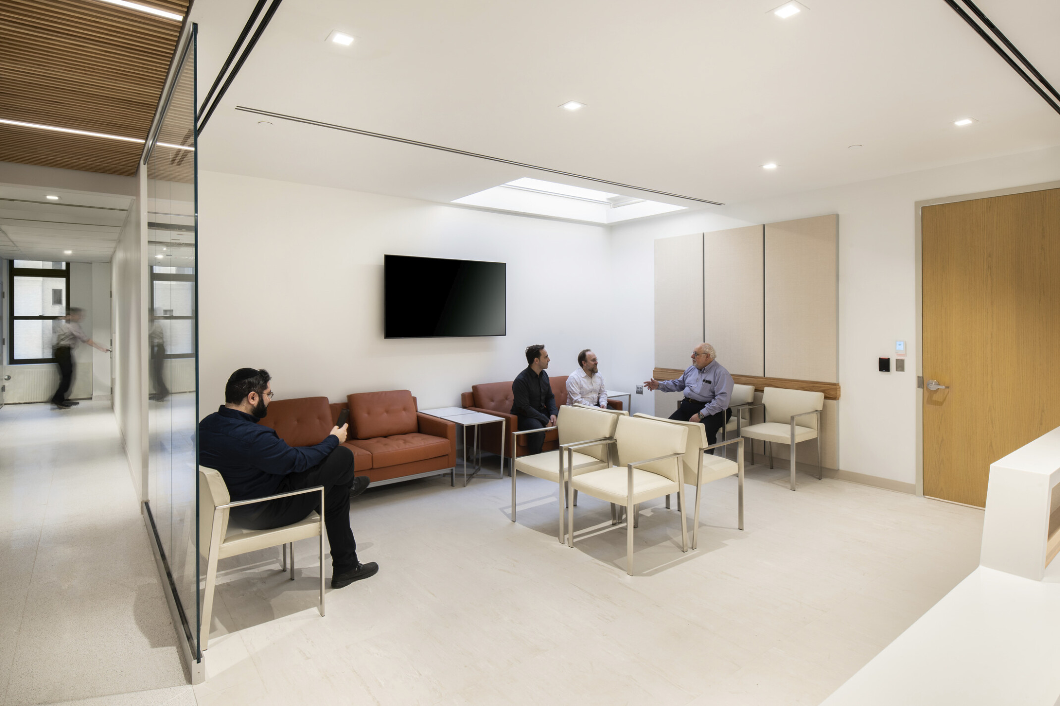 Group of individuals sitting in a hospital waiting area with off-white walls, a wooden door, off-white chairs and orange couches