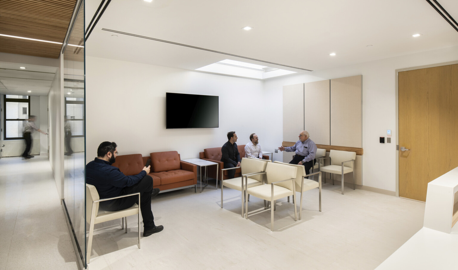 waiting room with a warm and restrained material palette with terracotta and white oak wood accents helps to reduce anxiety, a skylight brings natural light indoors, beige carpet, cream colored walls and ceiling lend a soothing sense of calm for patients