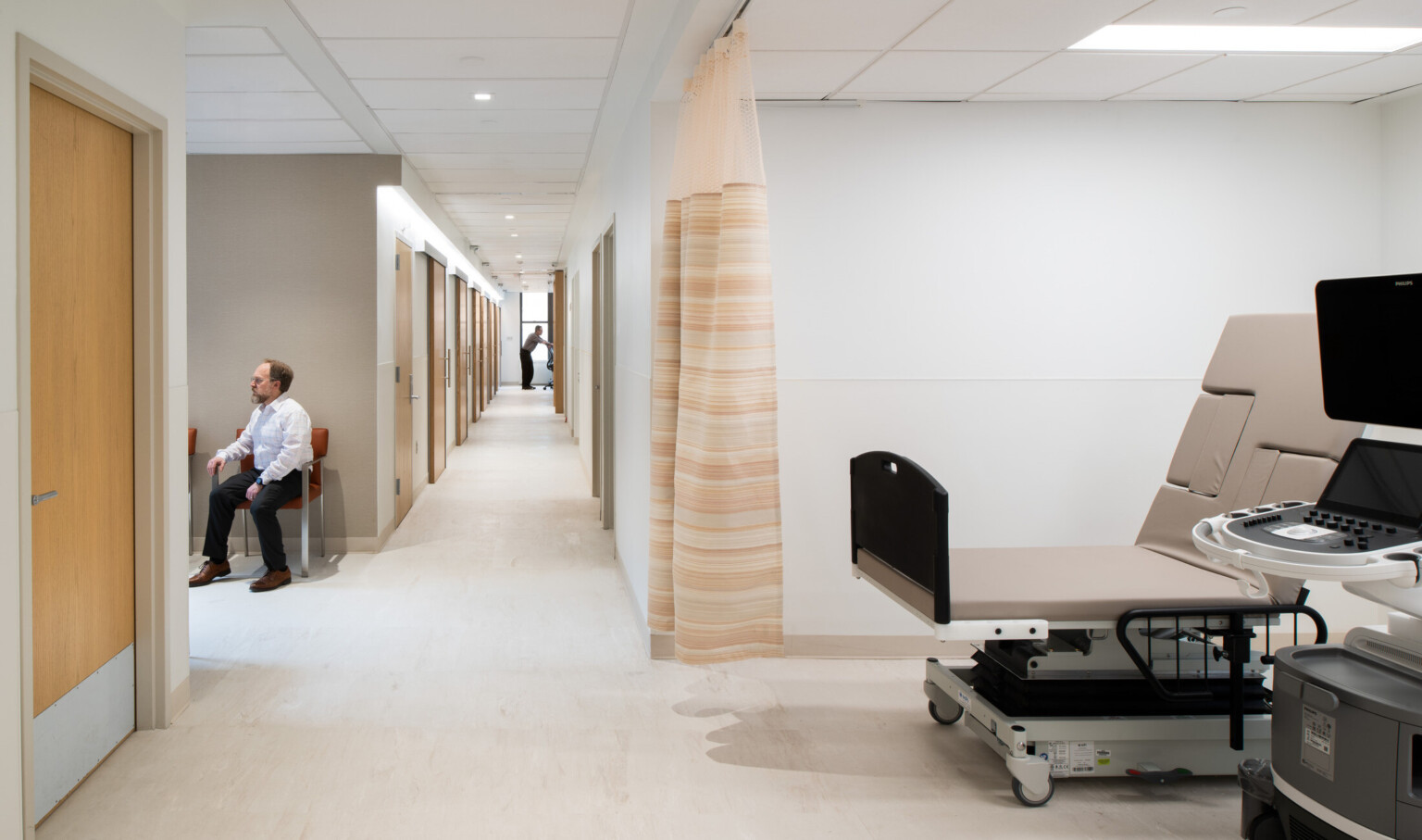 Medical office hallway with an exam room area on right with exam table and equipment. Man sitting in a waiting area in a red chair