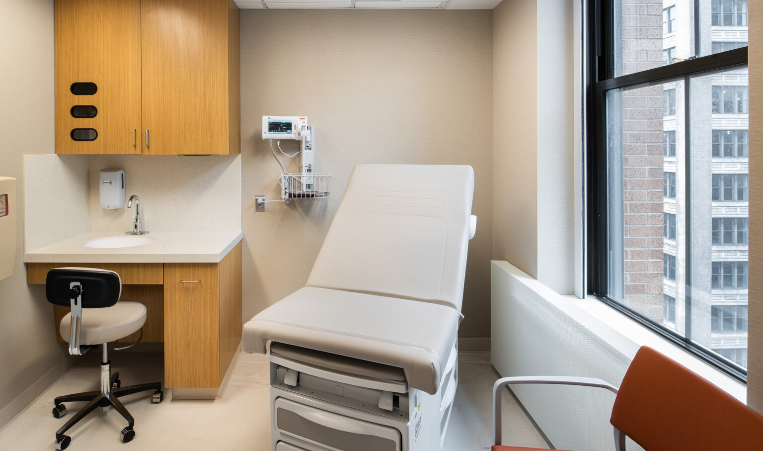Medical exam room with wooden cabinets, large black frame window, exam table, red chair and beige walls