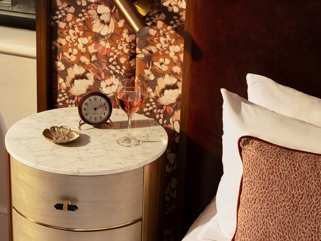 Nightstand at The Benjamin Royal Sonesta Hotel with a glass of wine, clock, and jewelry dish; headboard features a floral pattern