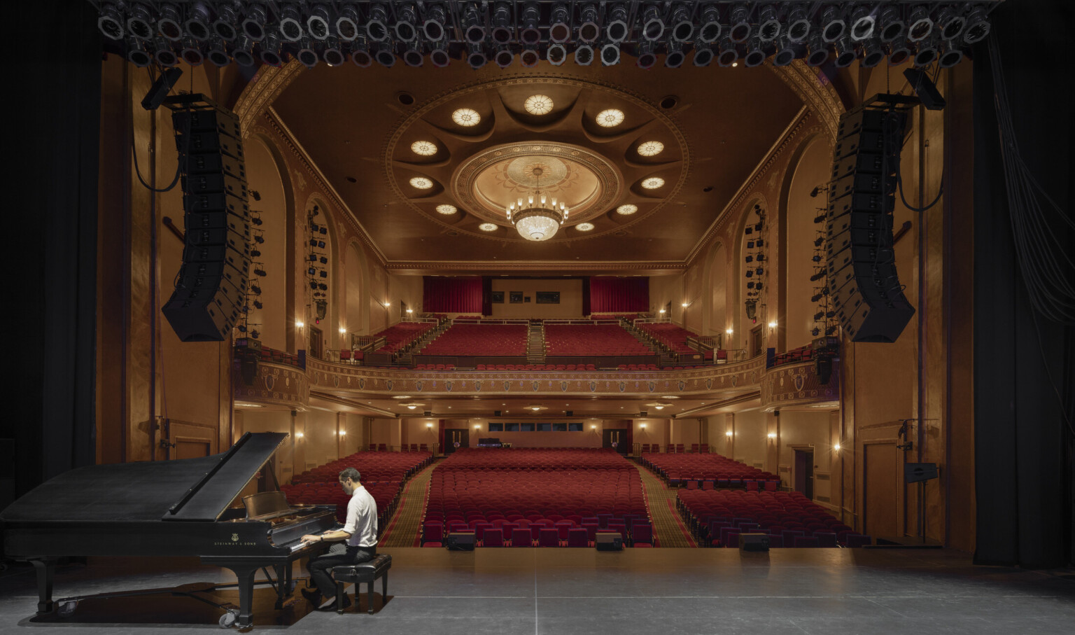 Theater from stage looking to red audience seats with large balcony. Round lighting details on ceiling encircling chandelier