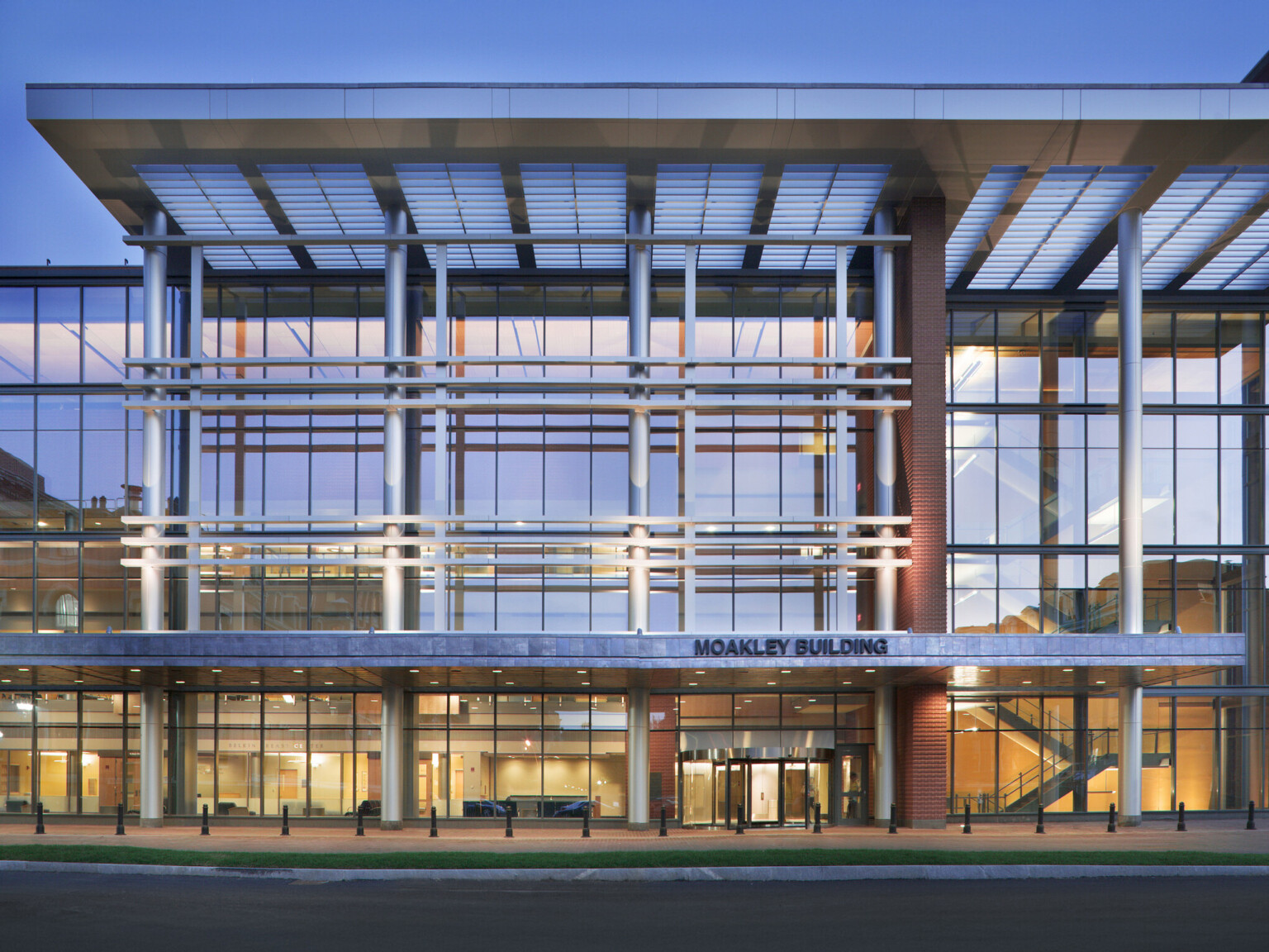 Exterior view of a modern medical building with large glass windows and a white grid-like façade. The interior is brightly lit, showing staircases and open spaces.