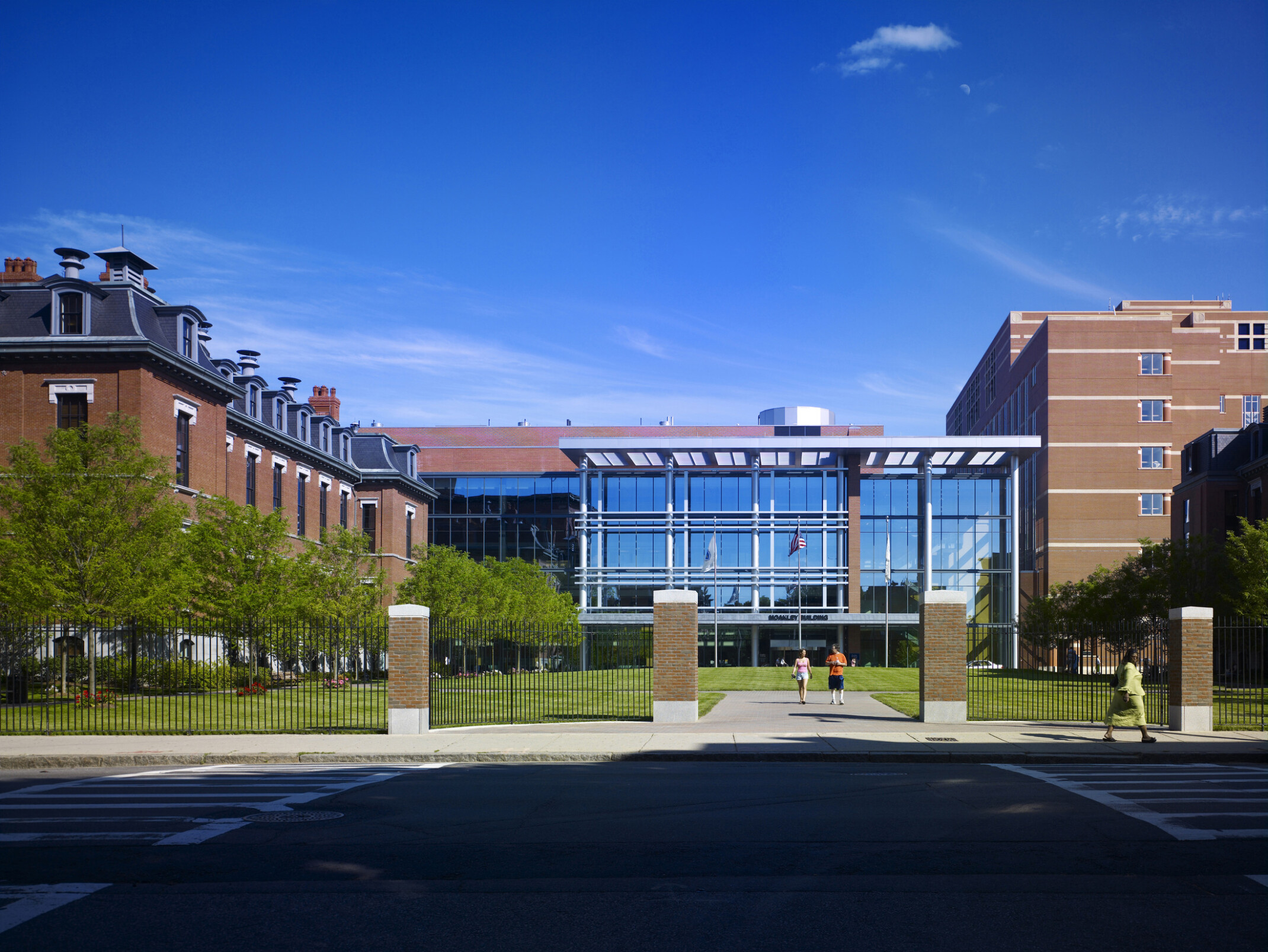 Exterior of a modern glass building between two brick buildings. A gated path leads to the entrance.